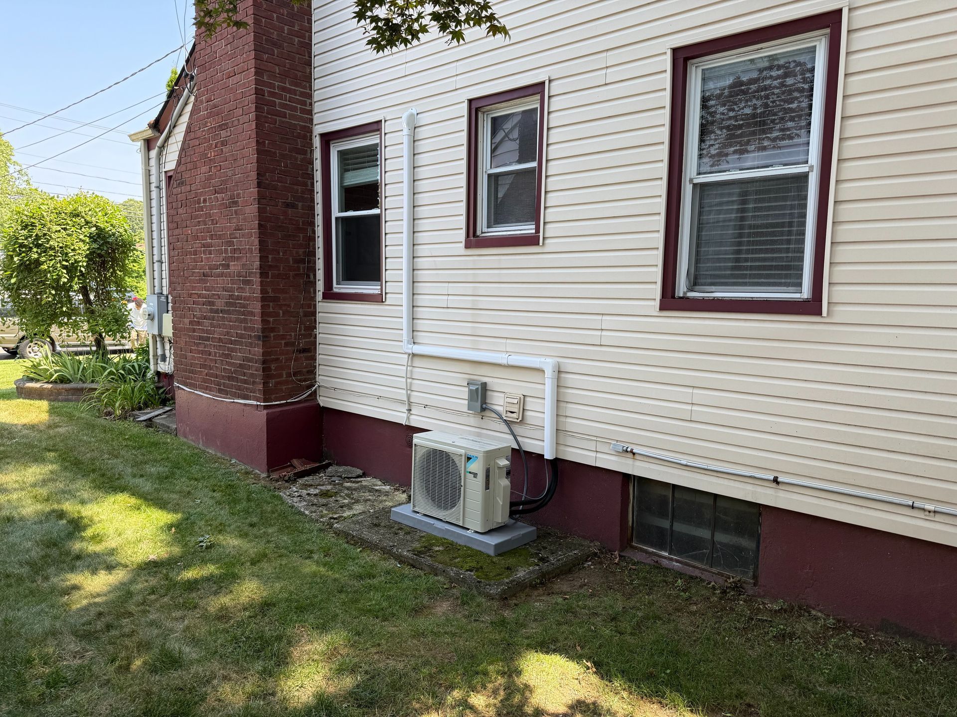 Side of a beige house with burgundy trim and a brick chimney, an outdoor AC unit, and windows.