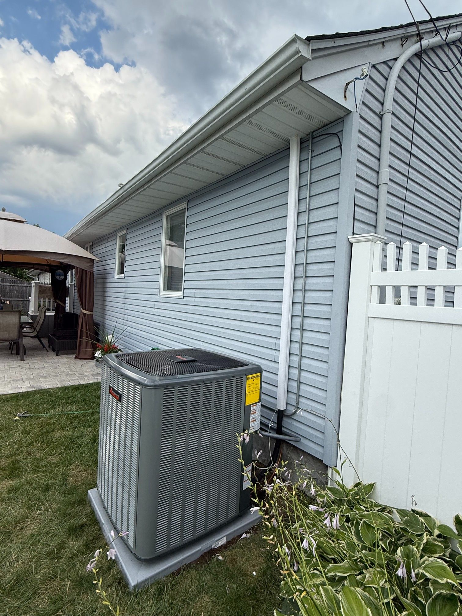 Air conditioning unit outside a light blue sided house next to a white fence and grass.