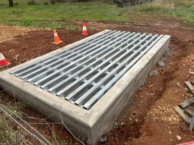 Metal cattle guard installed in a concrete base on dirt, with orange safety cones in the background.