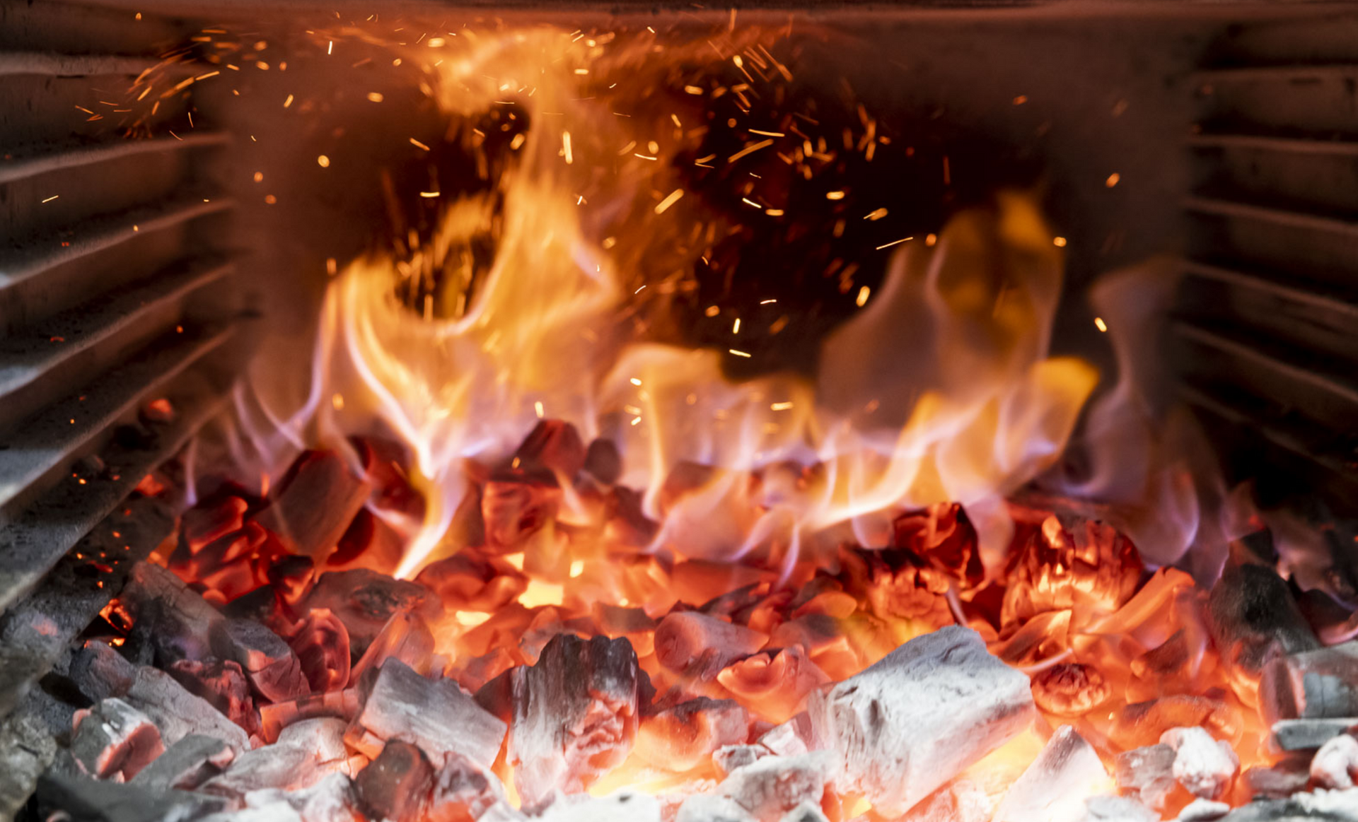 Des flammes engloutissent du charbon de bois incandescent dans un four, avec des étincelles et de la fumée qui s'élèvent.
