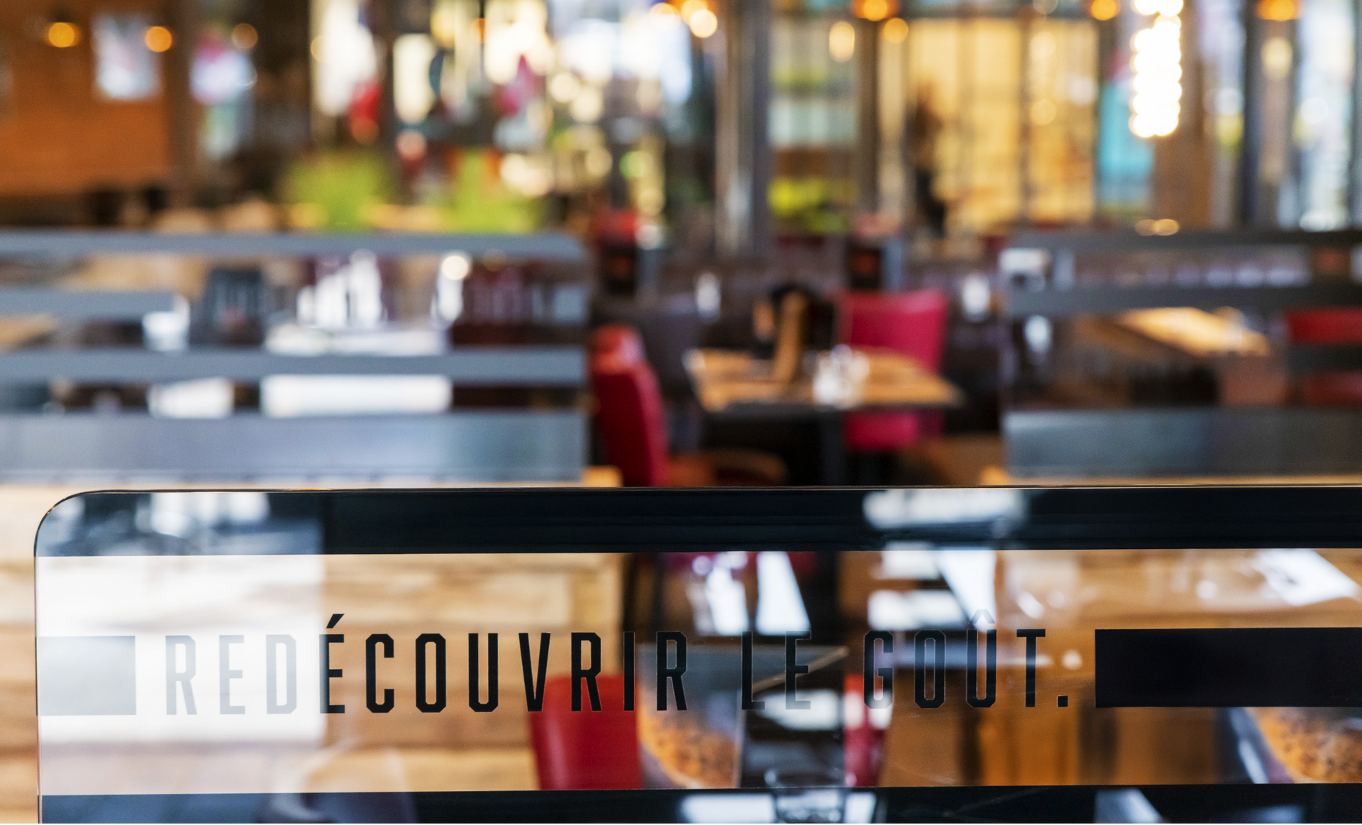 Intérieur du restaurant : arrière-plan flou avec tables et chaises. Texte sur verre : « REDÉCOUVRIR LE GOÛT ».