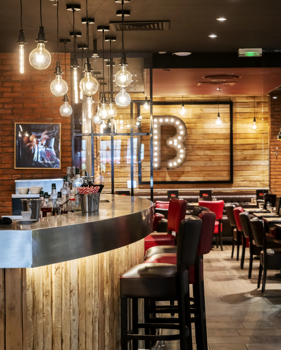 Intérieur de restaurant avec bar, tabourets, tables, sièges rouges, briques apparentes et une lettre B éclairée.
