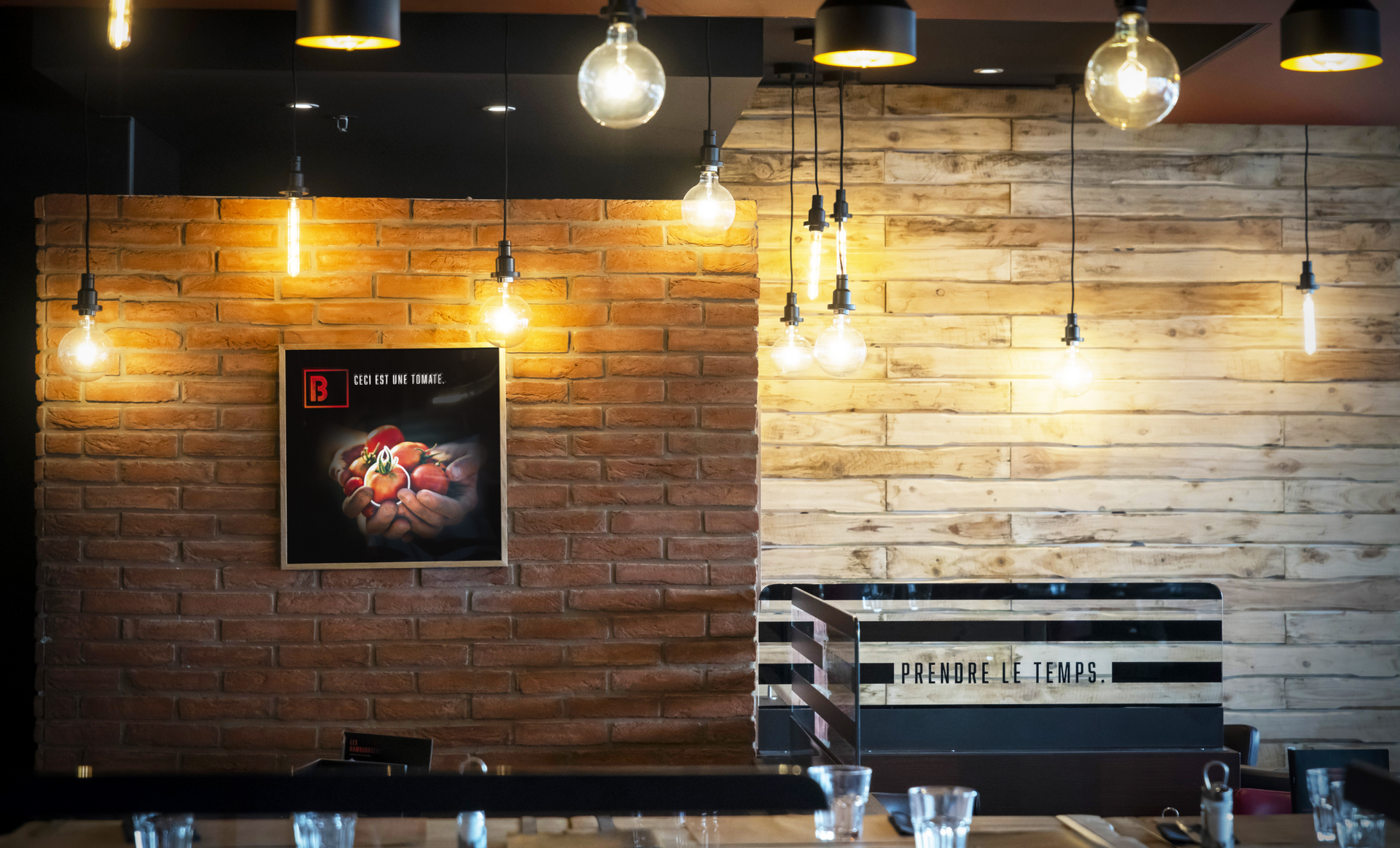 Intérieur de restaurant avec murs en briques et en bois, lumières suspendues et œuvre d'art encadrée.