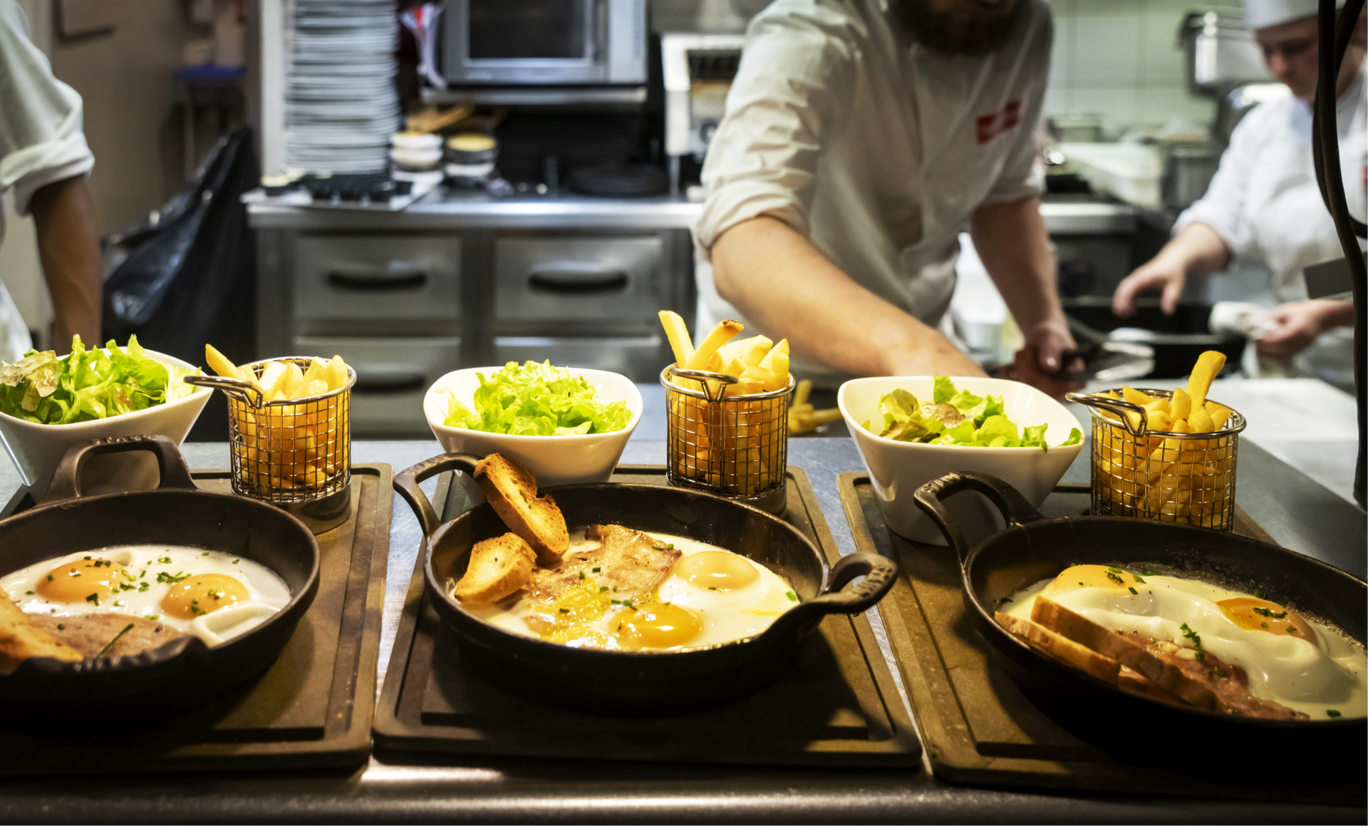 Plats cuisinés sur le comptoir, chefs en arrière-plan. Poêlées avec œufs, frites et légumes en accompagnement.