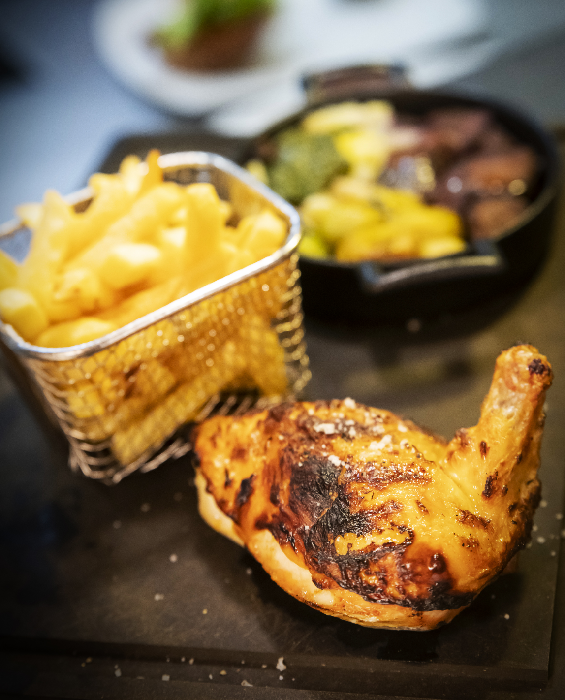 Cuisse de poulet rôtie, frites dans un panier et légumes sautés sur une surface en bois foncé.
