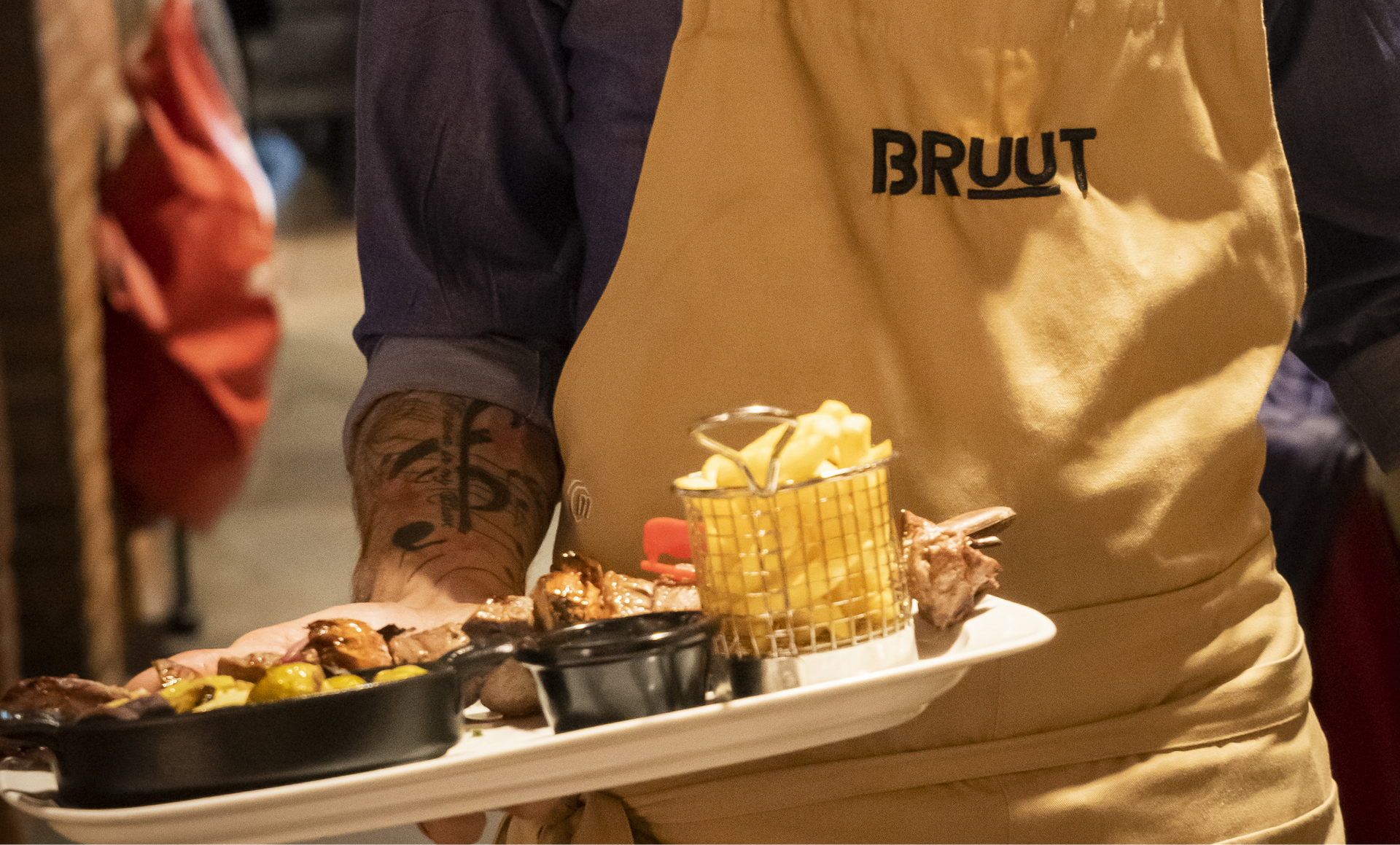 Personne portant un tablier tenant un plateau de nourriture : steak, frites et autres plats. Le logo « BRUUT » est visible.