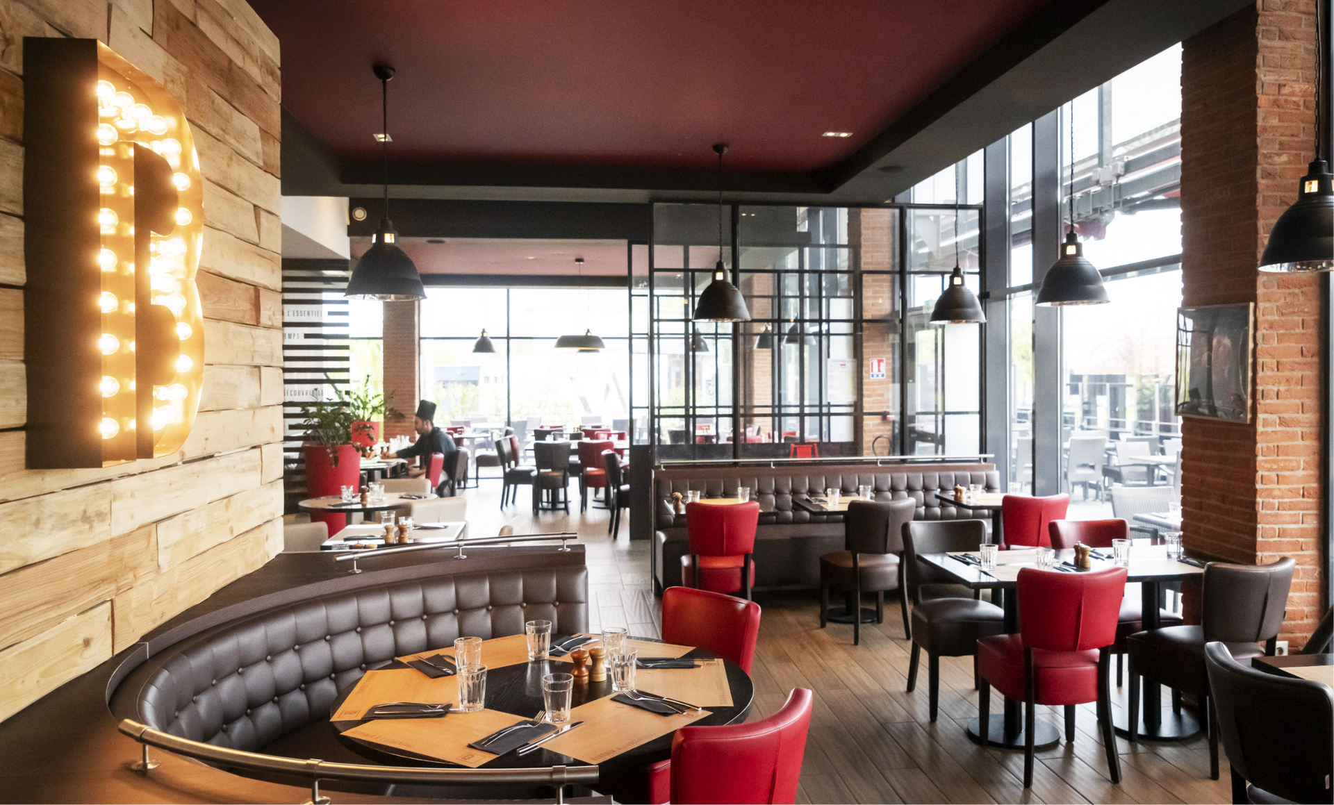 Intérieur de restaurant avec sièges rouges et noirs, briques apparentes et grandes fenêtres.