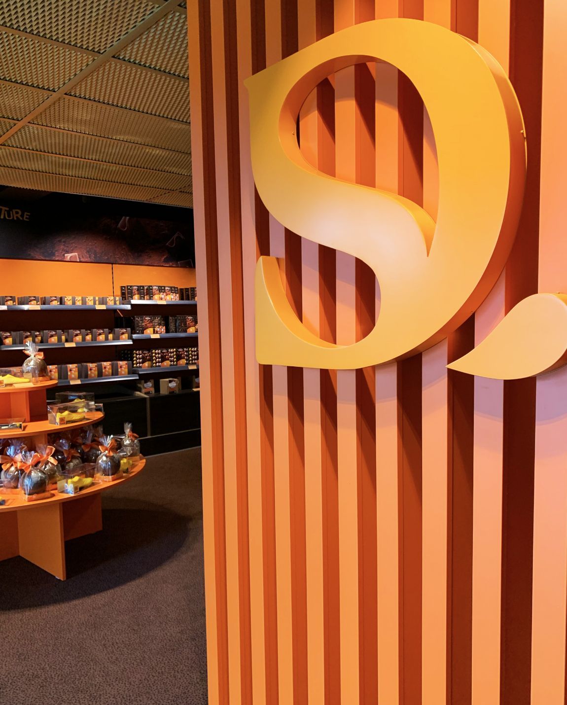Panneau avec un « S » stylisé sur un mur à rayures orange, porte d'entrée vers un magasin avec étagères et présentoirs.