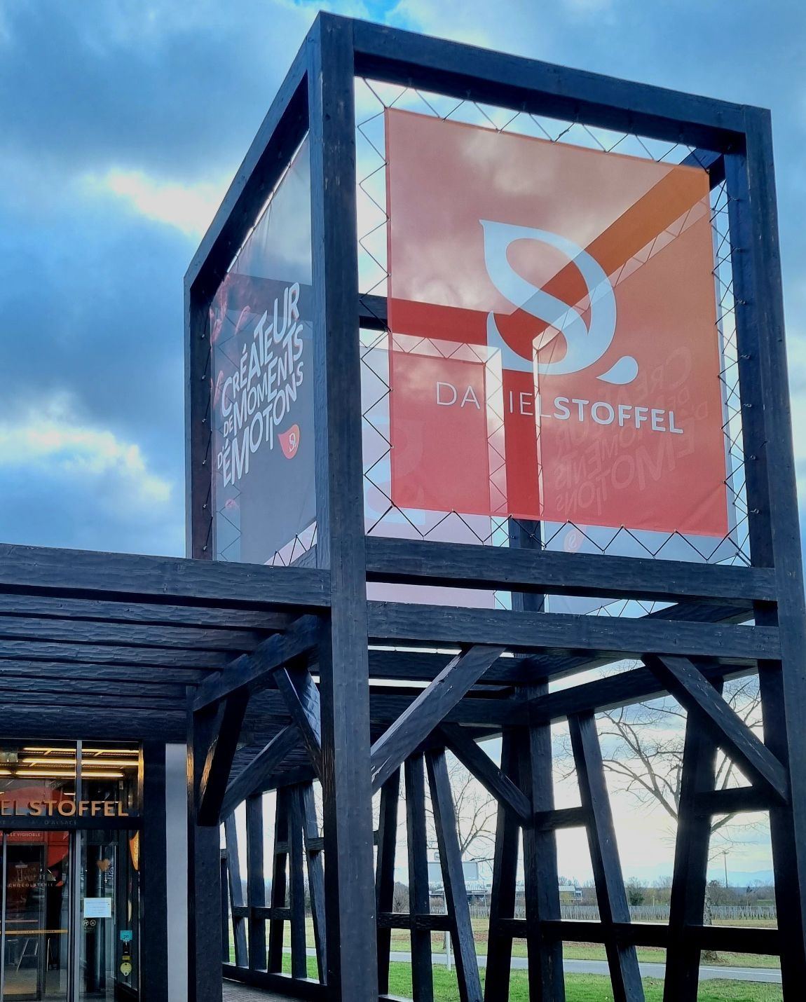 Structure en bois noir avec panneau orange et blanc « ISS. D'STOFFEL ». Ciel nuageux en arrière-plan.