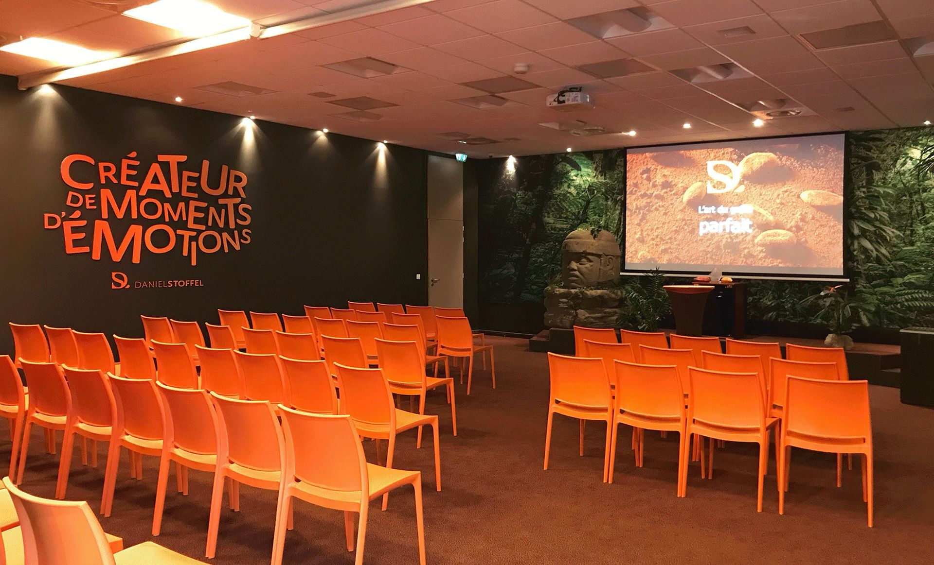 Salle de conférence avec chaises orange et écran. Murs sombres avec texte.