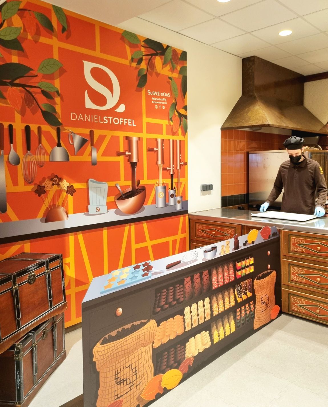 L'intérieur d'une chocolaterie avec une personne portant un masque ; art mural orange, étagères de produits.