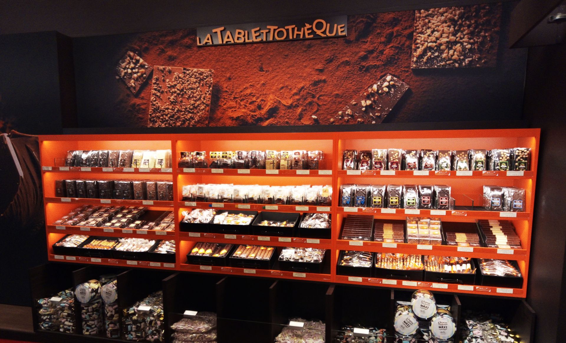 Présentoir de chocolaterie avec étagères orange, rempli de divers produits chocolatés, sous une enseigne marron et orange.