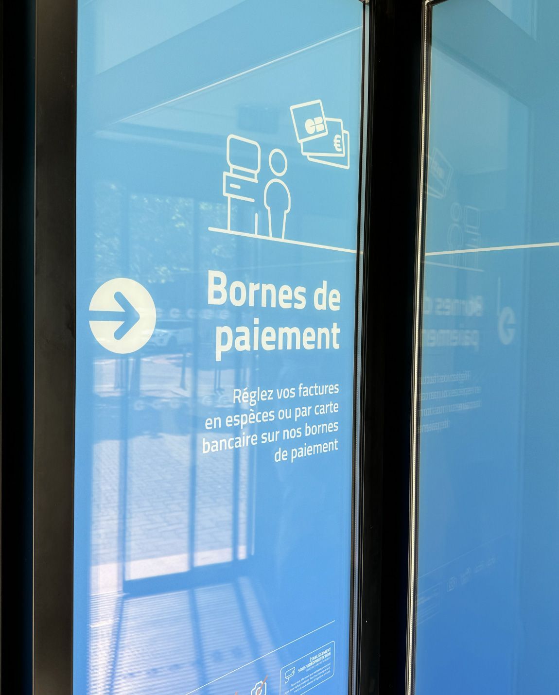 Signalétique indiquant les terminaux de paiement. Fond bleu, texte et icônes blancs. Comprend une flèche et une illustration d'une personne devant le terminal.