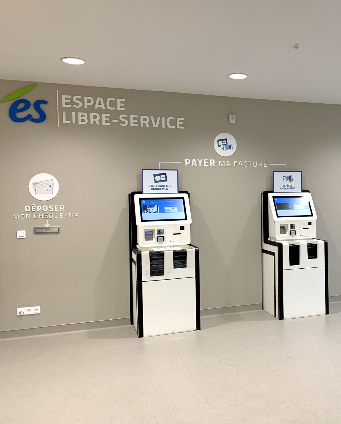 Deux bornes libre-service avec écrans, dans une pièce. L'inscription sur le mur indique « Espace Libre-Service ».