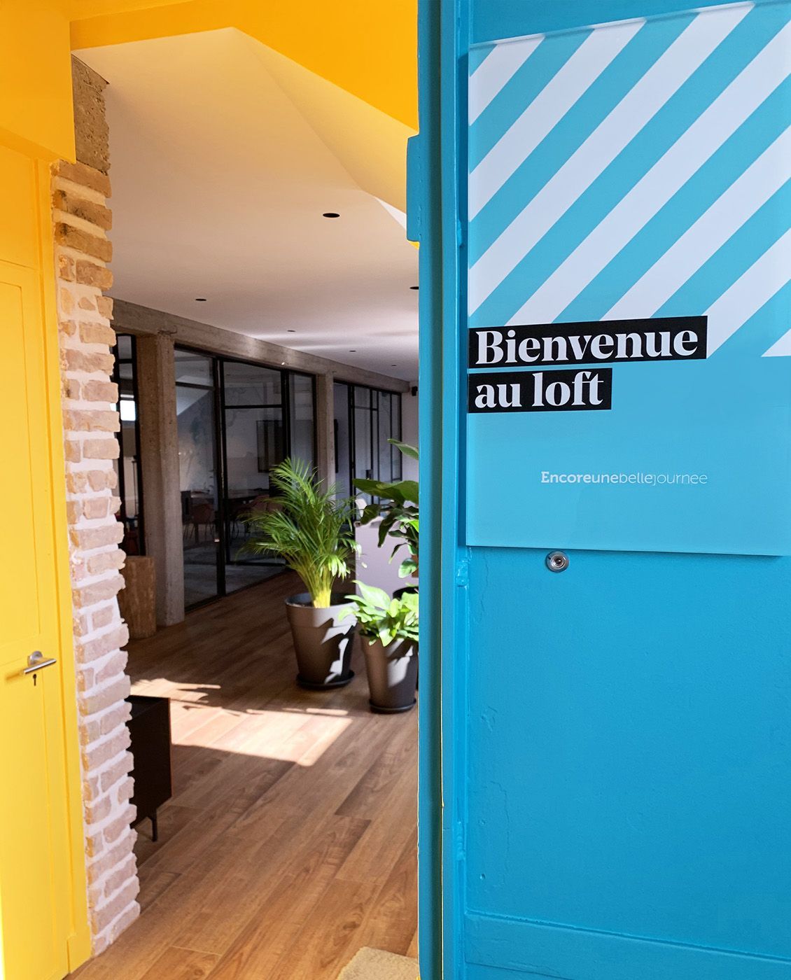 Porte bleue ouverte avec l'inscription « Bienvenue au loft ». Un mur jaune et des briques encadrent le couloir intérieur.