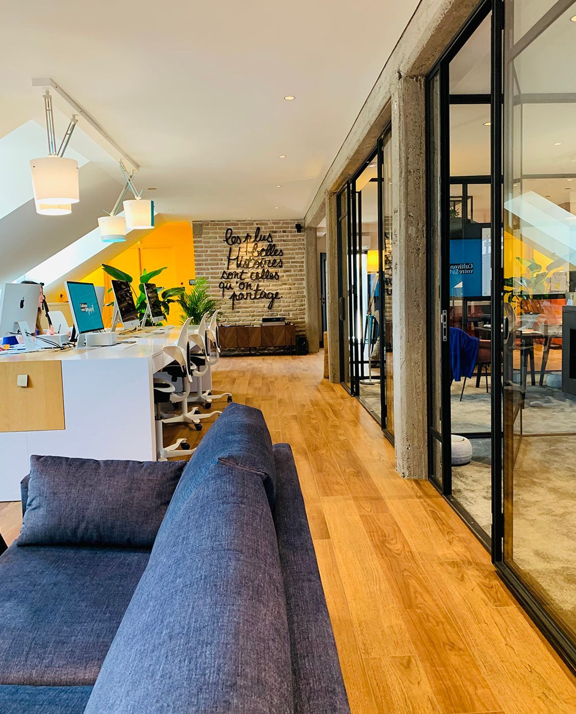 Intérieur de bureau : canapé bleu, parquet, bureaux aux parois vitrées, mur jaune et espaces de travail.