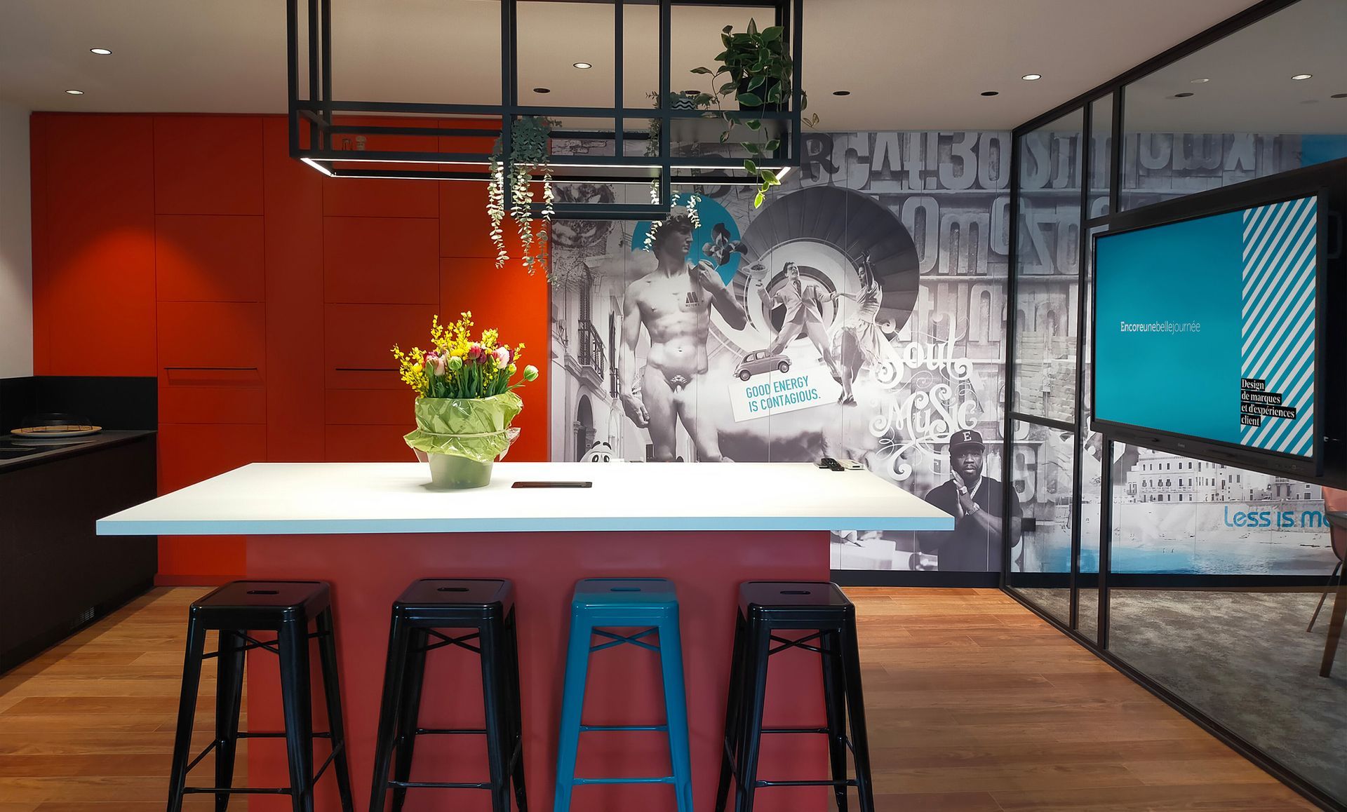 Cuisine avec îlot, tabourets et décoration murale, avec armoires rouges, luminaires noirs et grand écran.