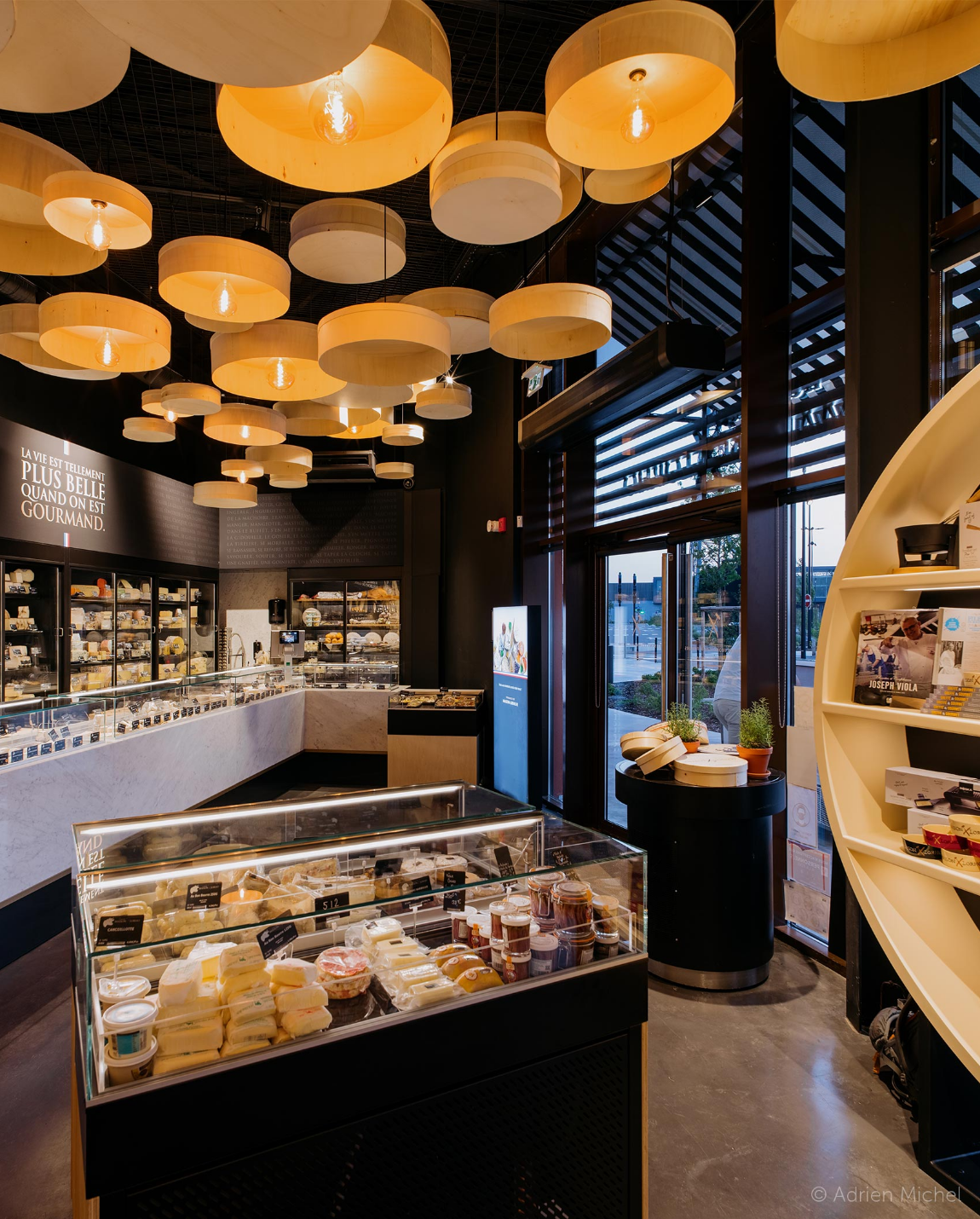 Intérieur de la fromagerie : vitrines avec fromage, globes lumineux suspendus, murs noirs et grandes fenêtres.