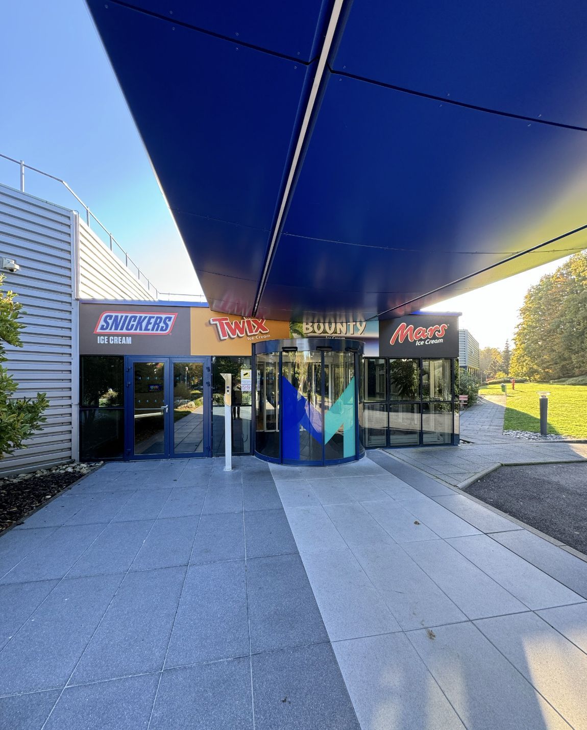 Entrée d'un centre commercial avec auvent bleu, magasins et portes tournantes.