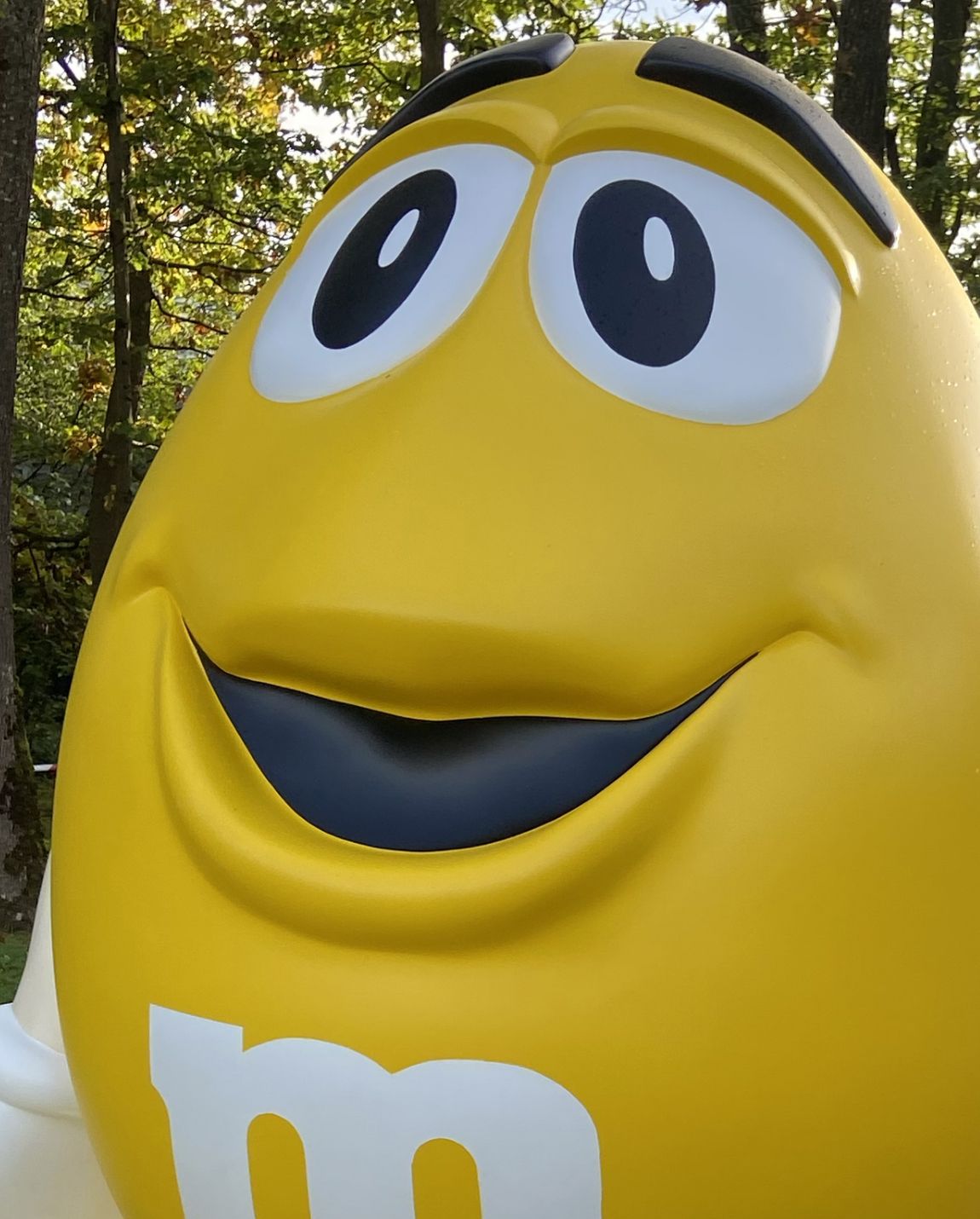 Personnage M&M jaune avec un visage large et souriant dans un décor de parc.