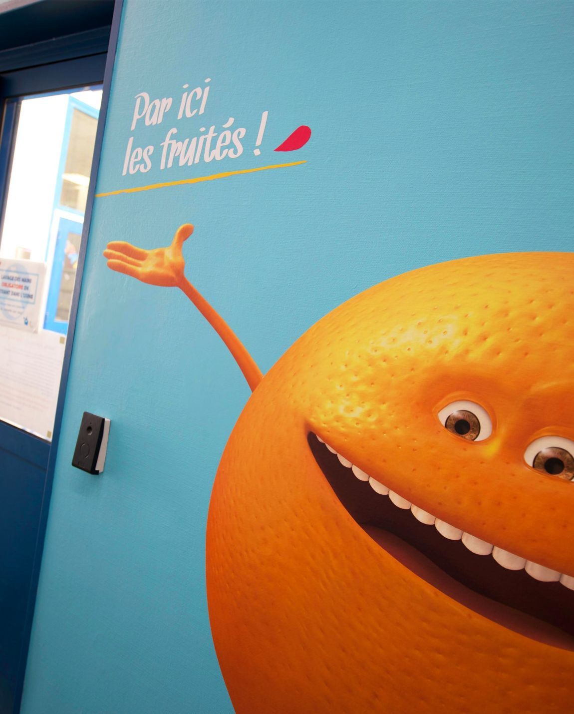 Un personnage de dessin animé orange avec un sourire et un bras levé pointe un panneau français au-dessus d'une porte.