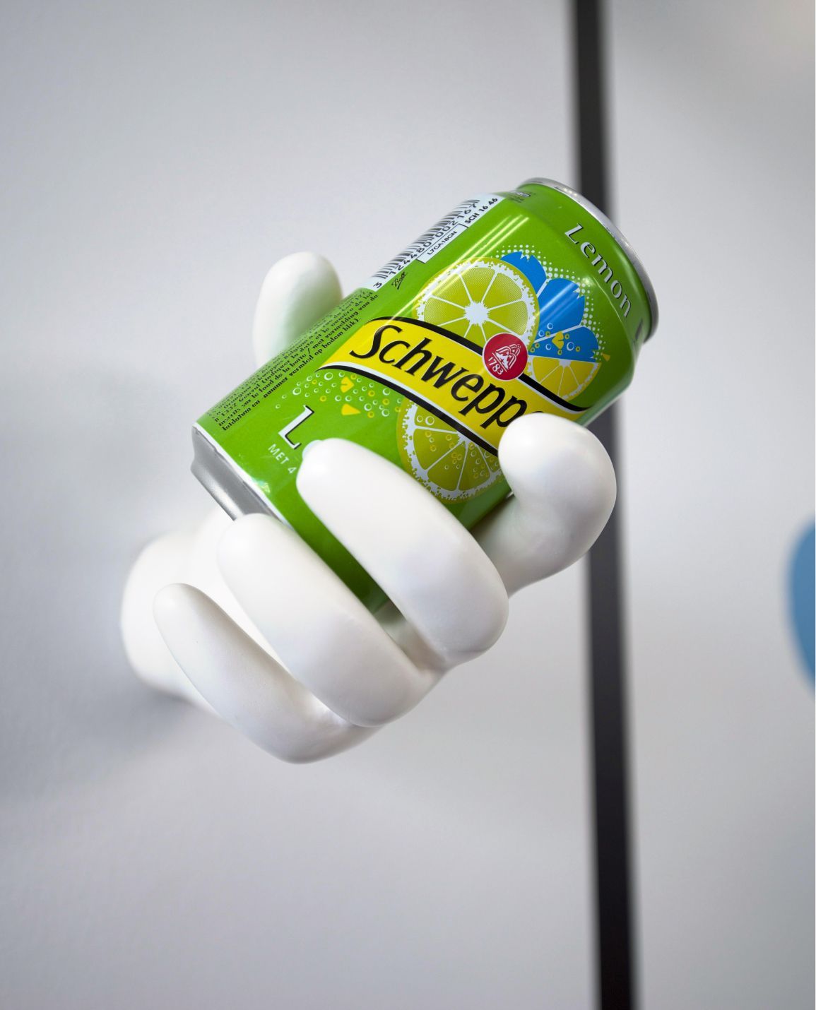 Sculpture de main blanche tenant une canette de Schweppes verte contre un mur de couleur claire.