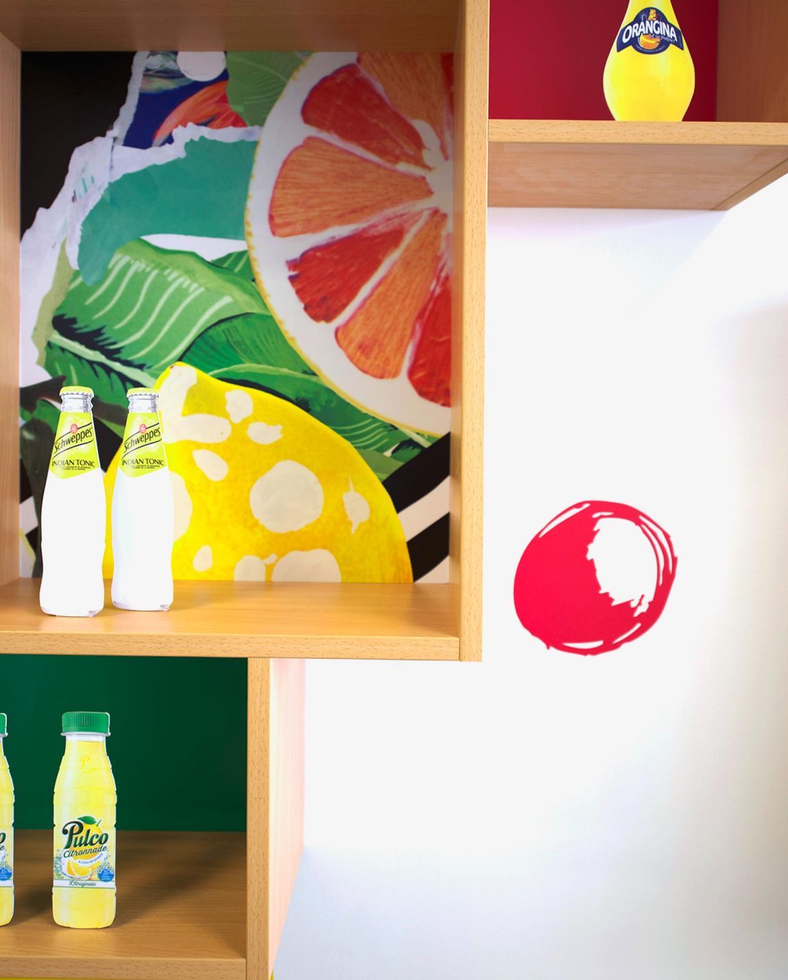 Des étagères en bois présentent des bouteilles de jus, un collage d'agrumes colorés et un cercle rouge sur un mur blanc.