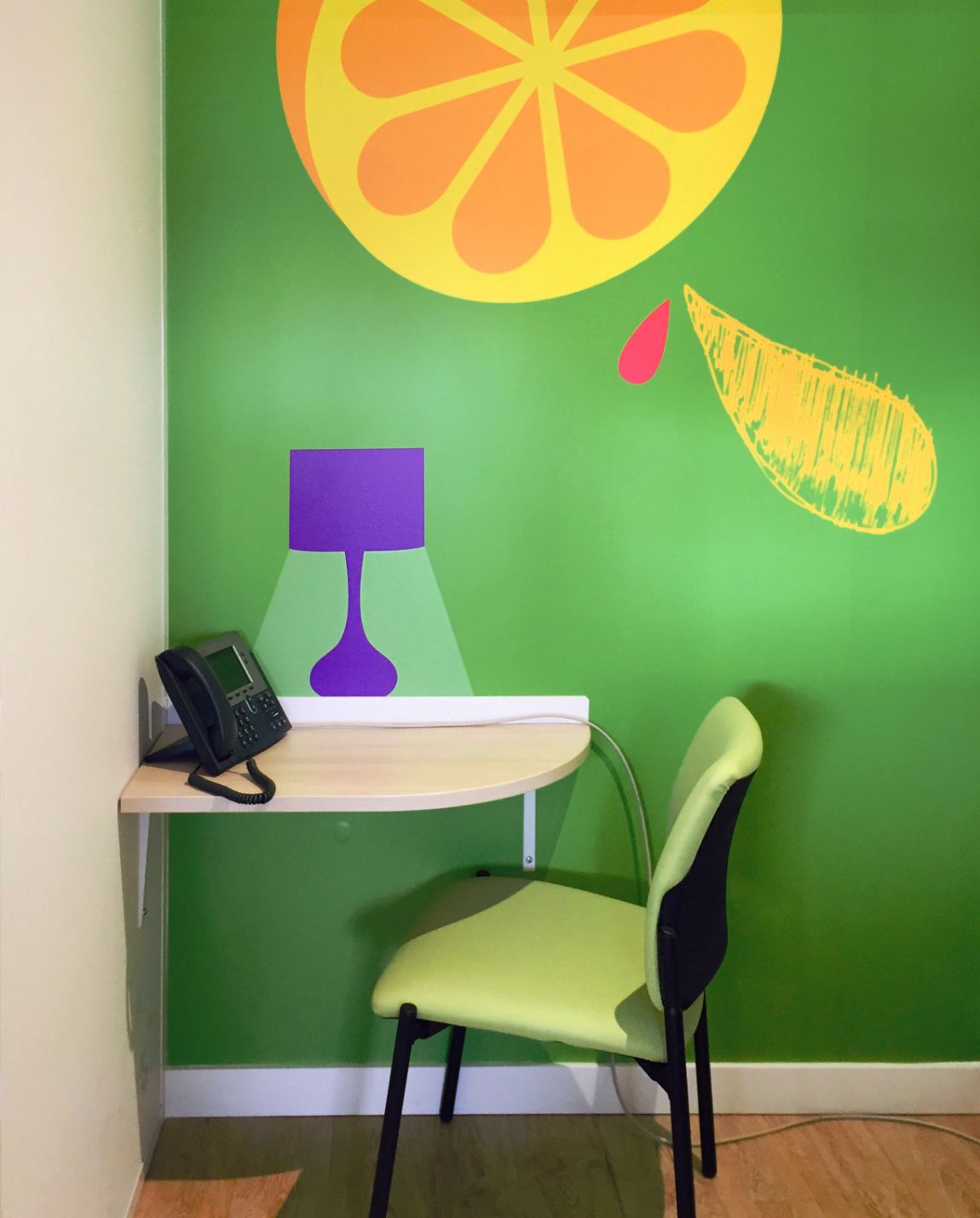 Mur vert avec une tranche d'orange murale, un petit bureau, une lampe violette, un téléphone noir et une chaise verte.