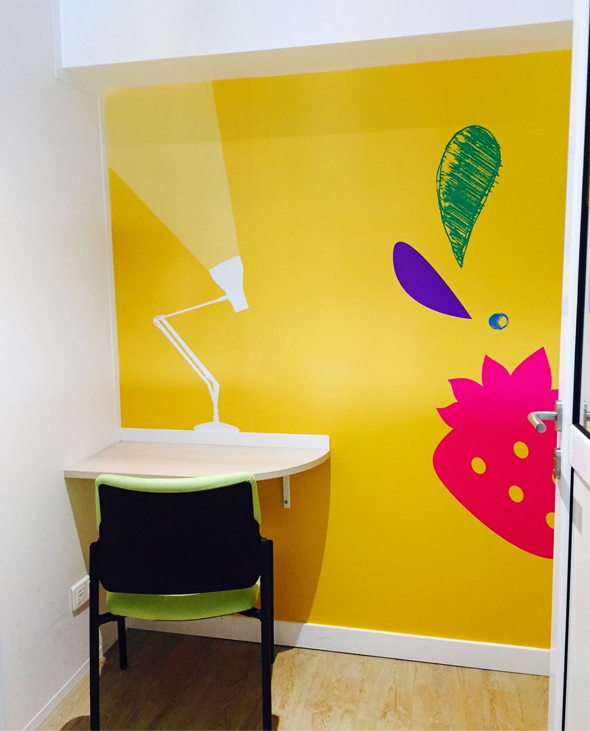 Un petit coin bureau avec un mur jaune, un bureau, une chaise et une peinture murale colorée sur le thème des fruits.