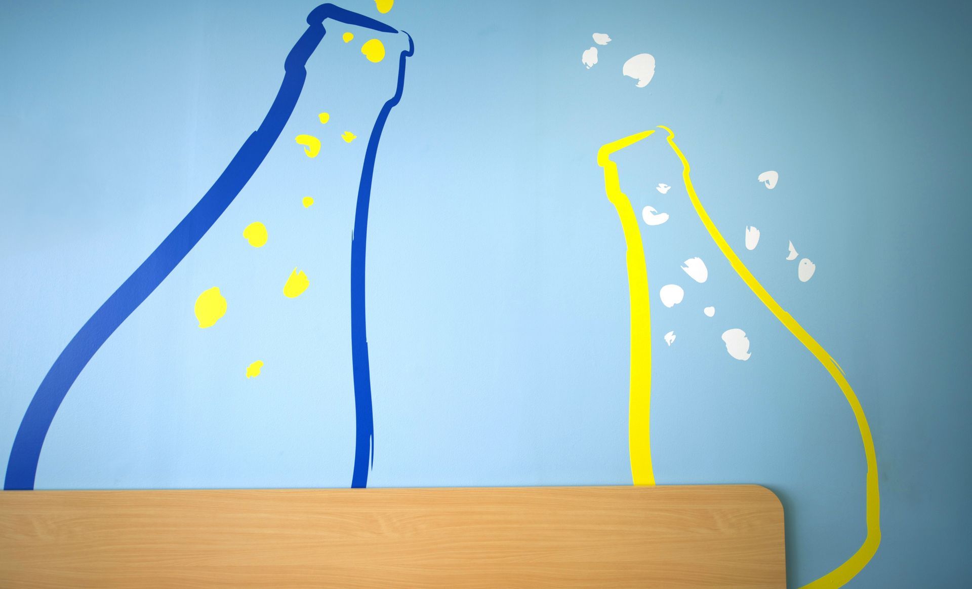 Bouteilles de champagne peintes en bleu et jaune éclatant sur un mur bleu ; table en bois au premier plan.
