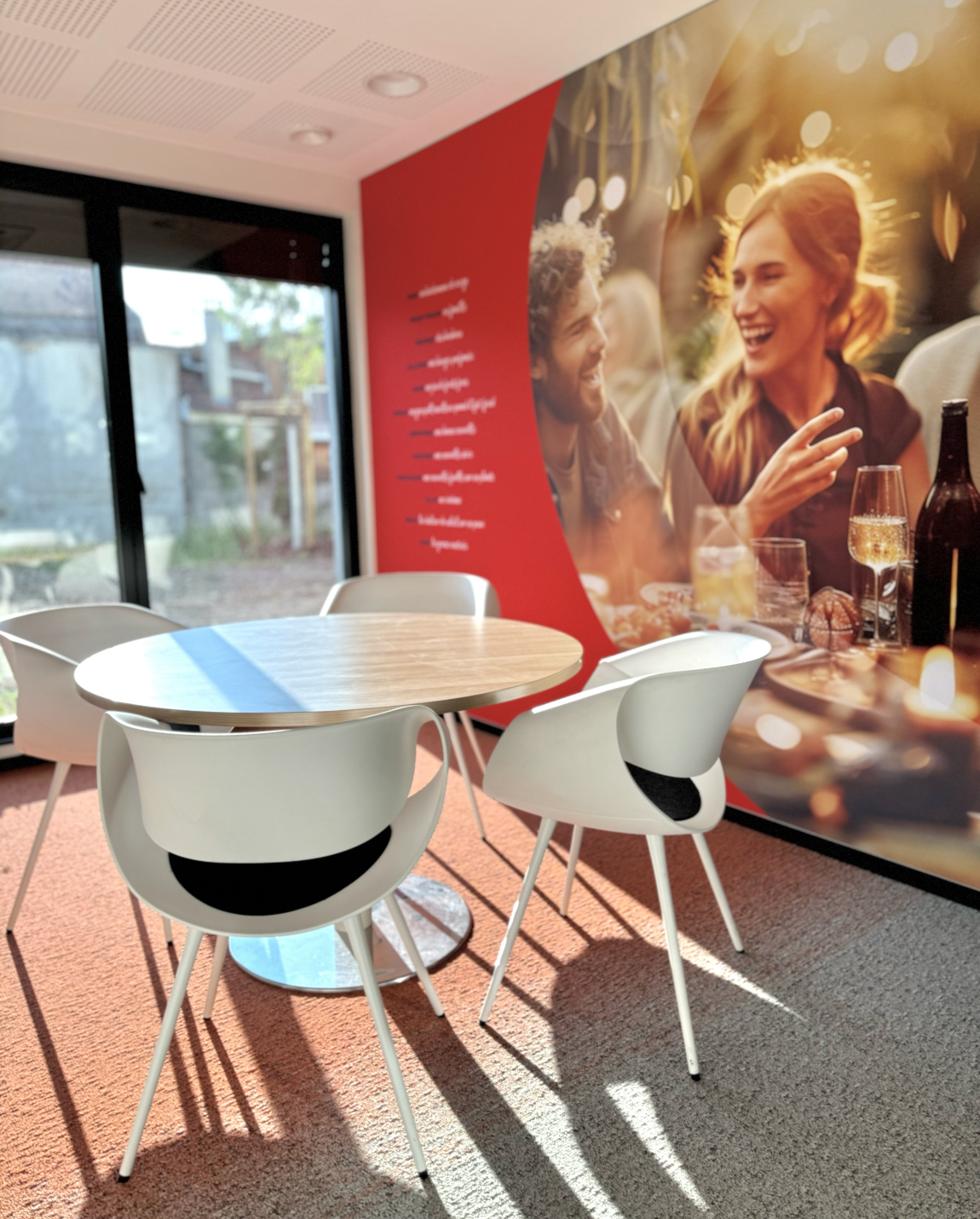 Table ronde avec chaises blanches, installée dans une pièce avec une fresque ensoleillée et un mur rouge.