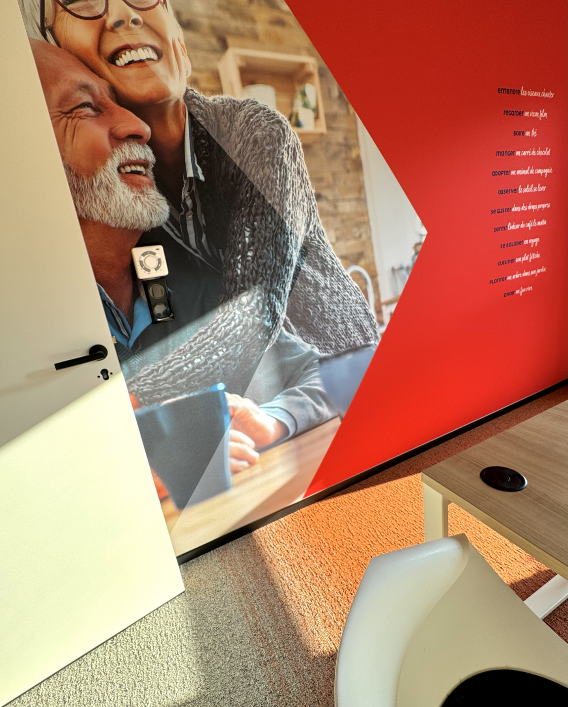 Couple de personnes âgées souriant, sur un mur. Coin rouge, table avec chaise et porte blanche.