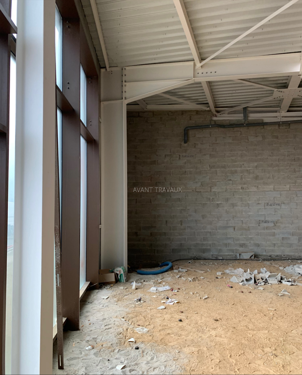 Vue de construction intérieure avec de grandes fenêtres, des poutres en acier apparentes et un mur de briques non fini.