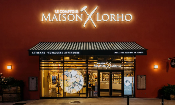 Boutique Maison Lorho avec enseigne lumineuse ; façade rouge, toit noir. Vitrines vitrées pour la présentation des produits.