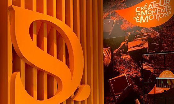 Grand logo orange « S » sur un mur strié ; à droite, un mur sombre avec le texte « CREATEUR DE MOMENTS D'EMOTIONS ».