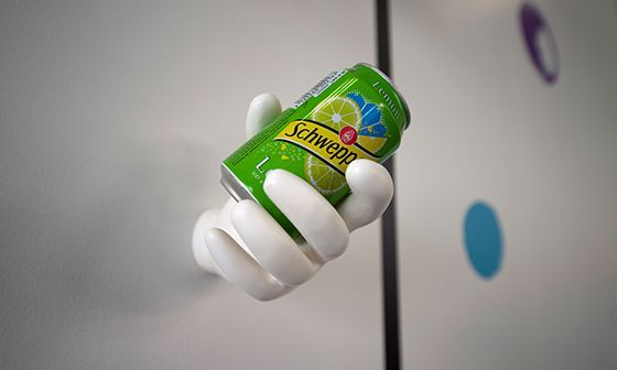 Sculpture de main blanche tenant une canette de Schweppes verte contre un mur blanc à pois colorés.