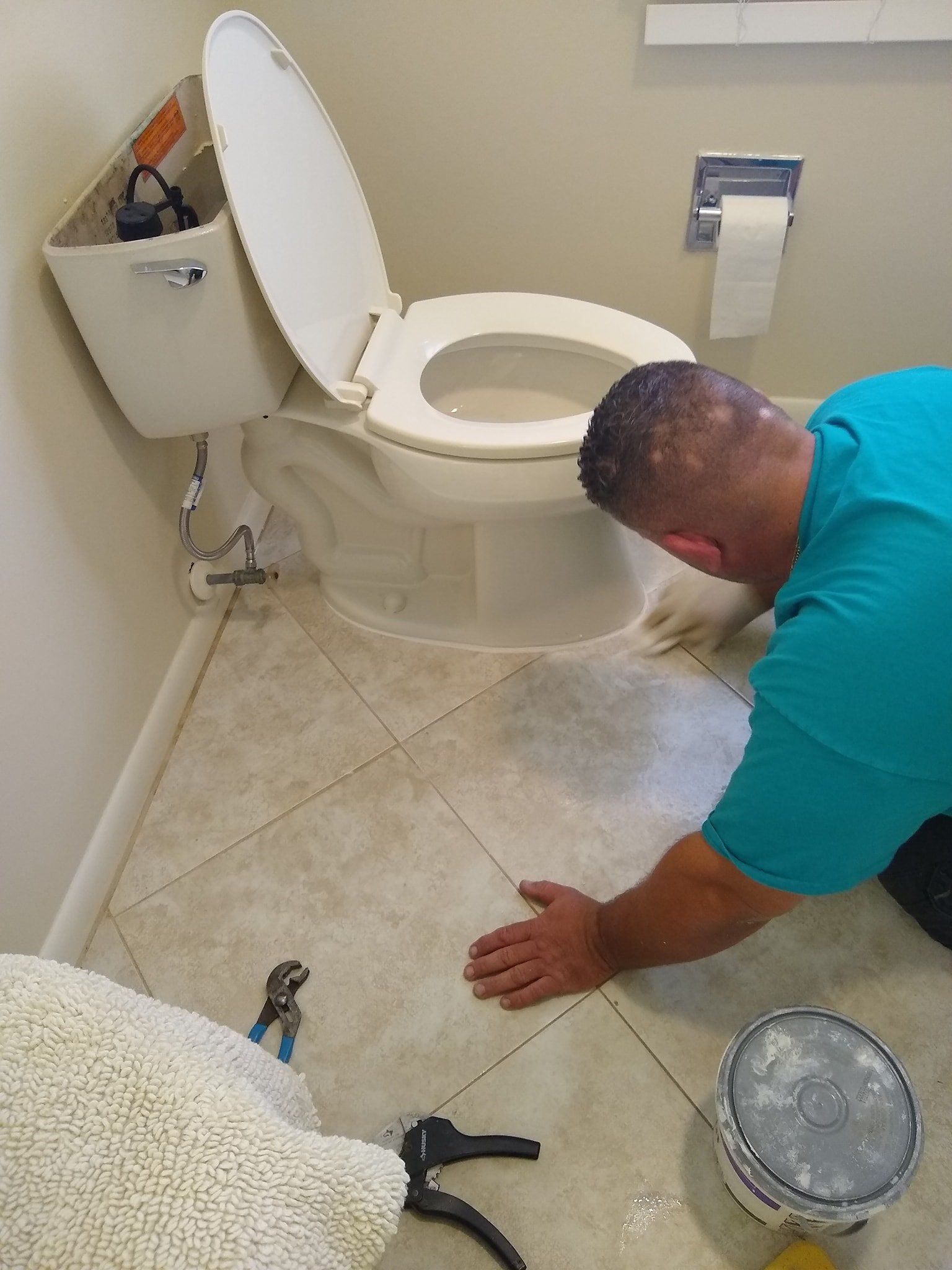 Man working on a toilet, kneeling on tiled floor. Toilet seat is up, parts exposed. Tools and bucket nearby.
