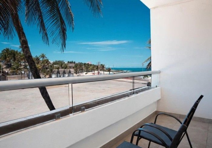 Balcón con vistas a una playa soleada y al océano azul, con una palmera y una silla en primer plano.