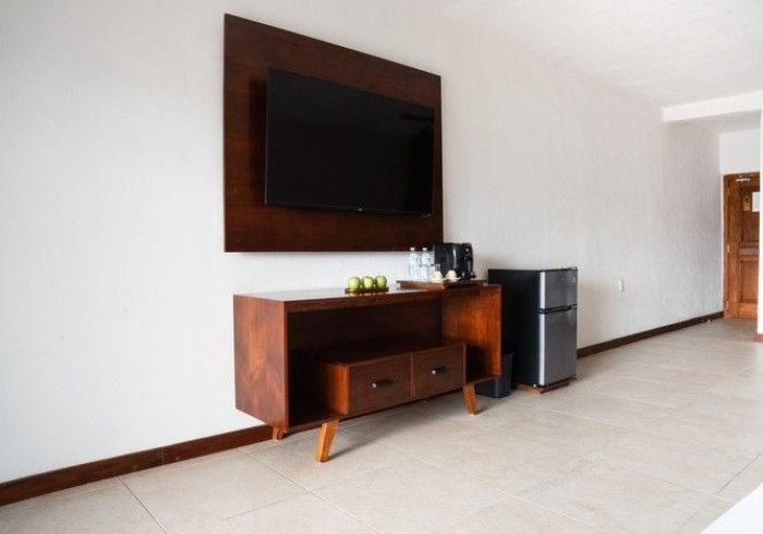 Habitación moderna con televisor montado en la pared, consola de madera y nevera pequeña sobre suelo de baldosas.