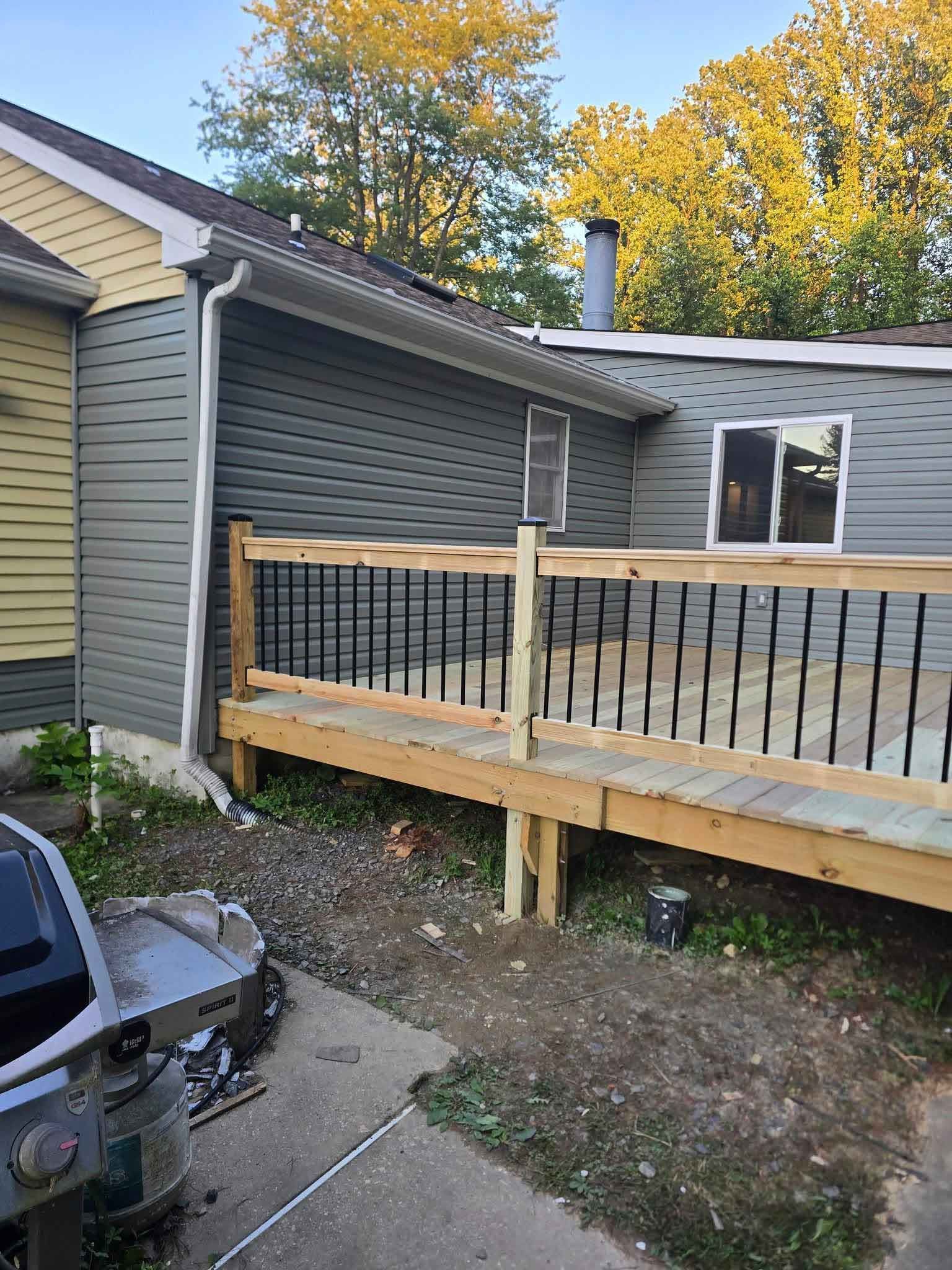 A wooden deck with black railings attached to a house with gray siding. The deck is unfinished, and a grill is in the foreground.