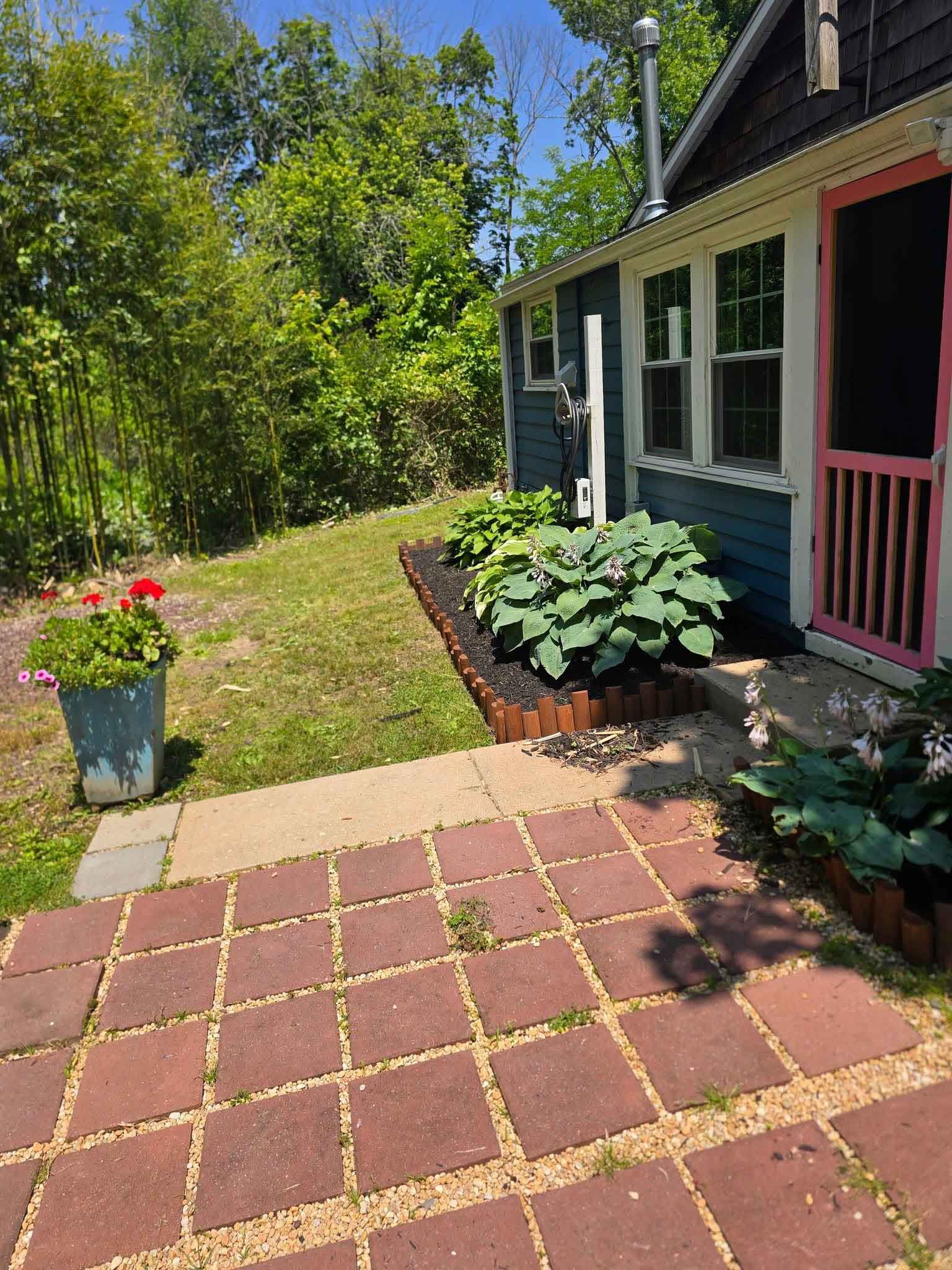 Brick patio leads to a blue house with pink doorframe and a flower garden. Potted flowers are to the left.