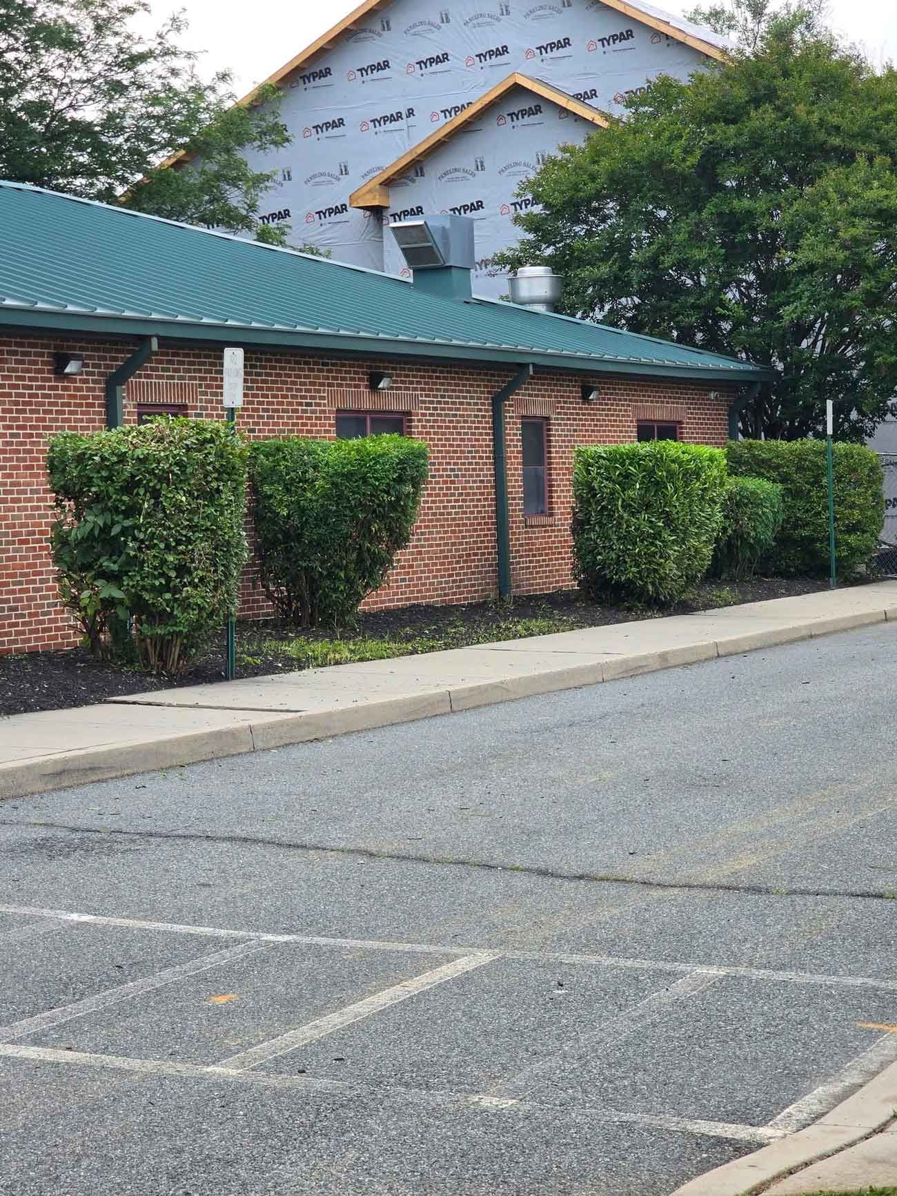 Brick building with a green metal roof, green bushes along the front, and a gray asphalt parking lot in the foreground. A building under construction is in the background.