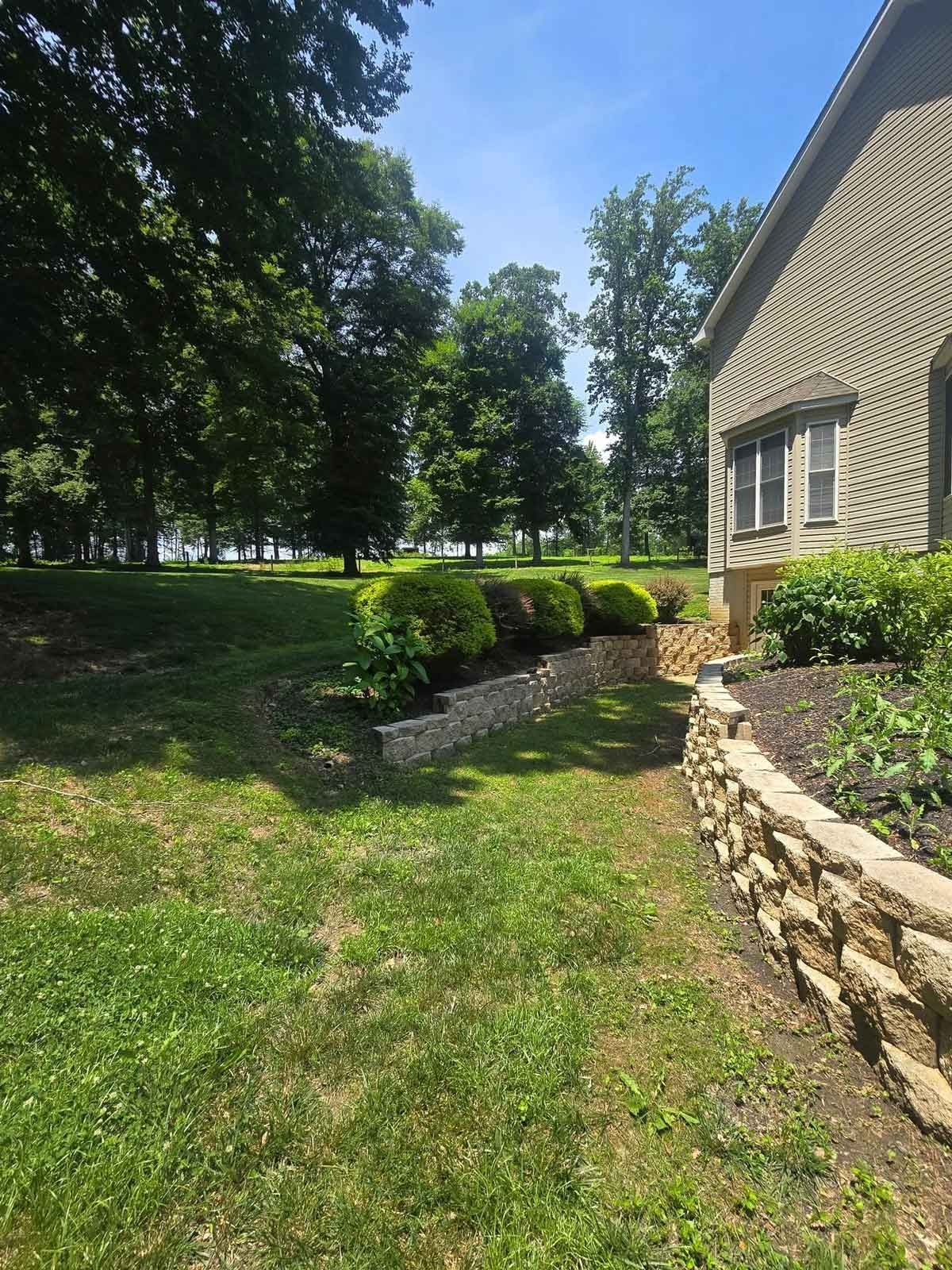 A grassy lawn slopes down to a tree-lined vista. A stone retaining wall runs along the side of a beige house.