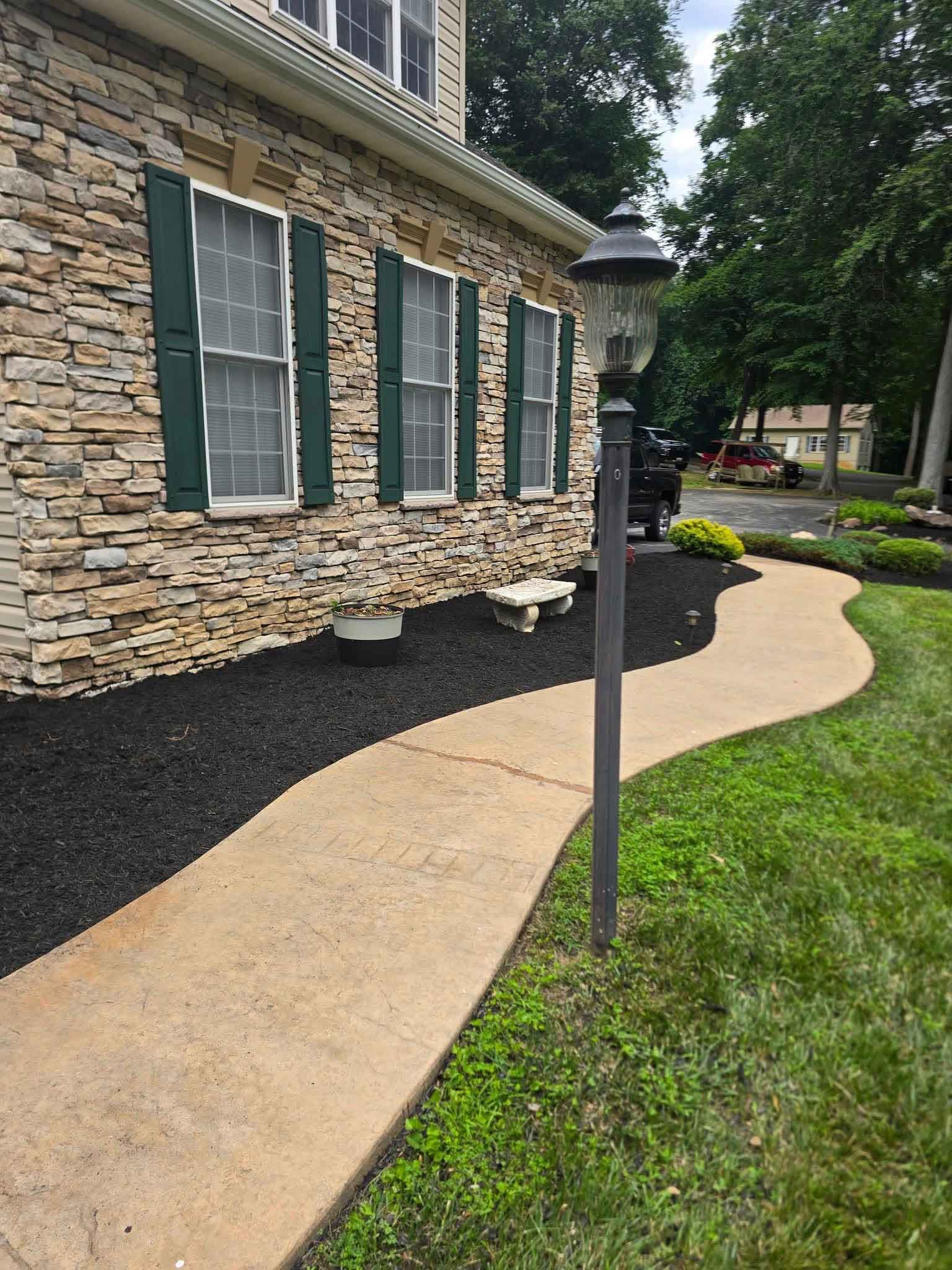 A stone-clad house with green shutters and a winding concrete path. A black mulch bed borders the path and the house.