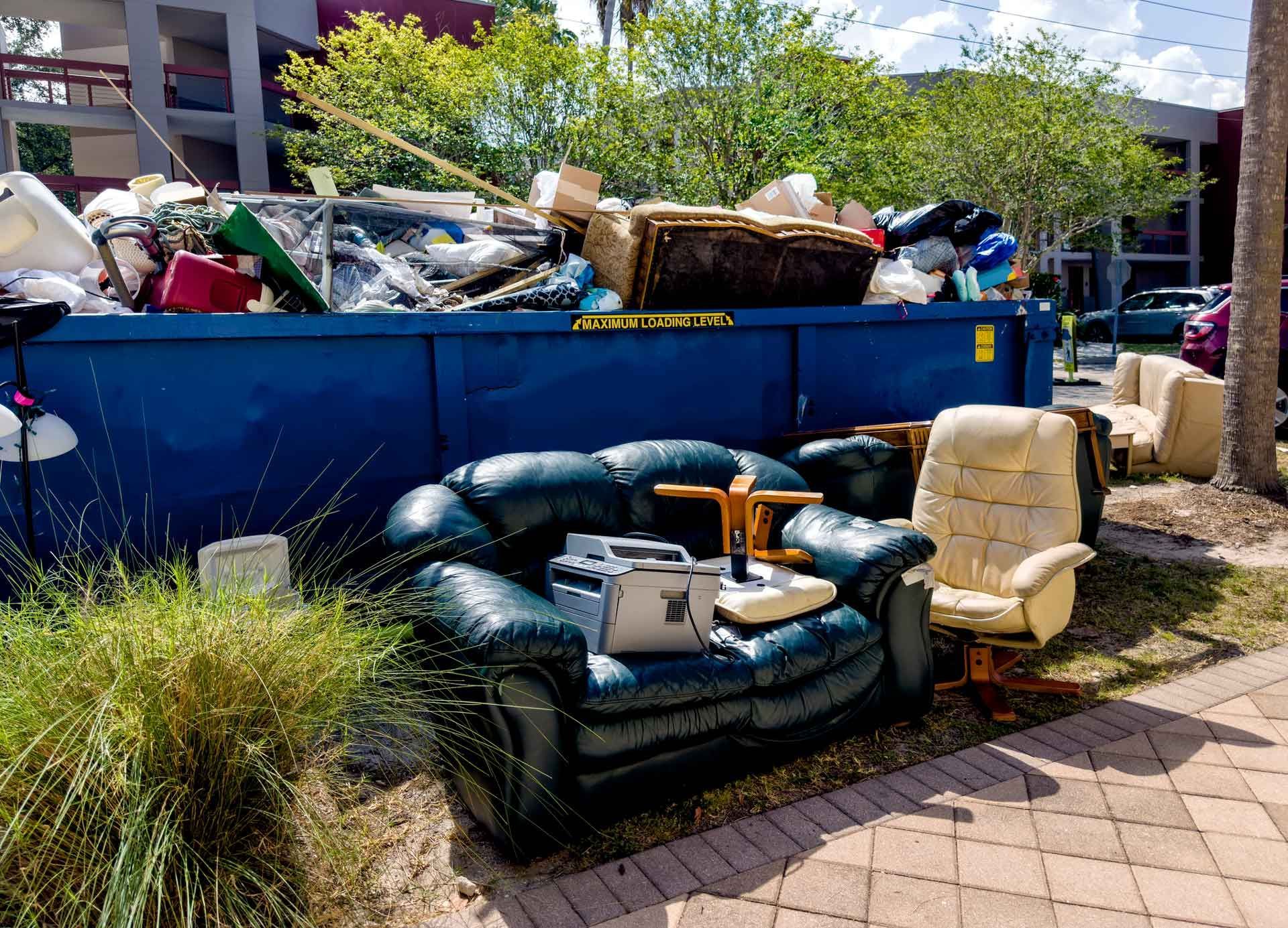 A full blue dumpster overflowing with trash next to discarded furniture like a green leather sofa and a beige recliner, outdoors on a brick patio.