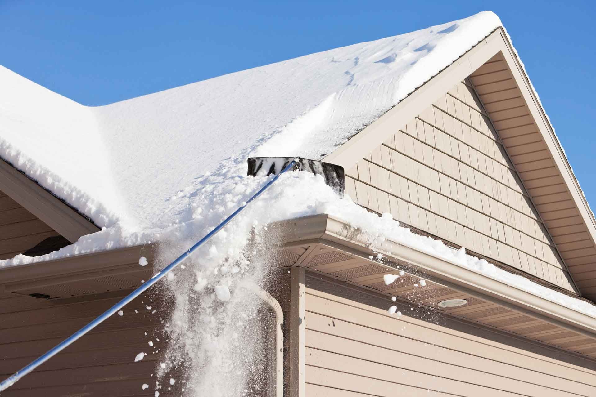 A person using a long-handled snow roof rake, clearing snow from a light-colored home's roof on a sunny day.