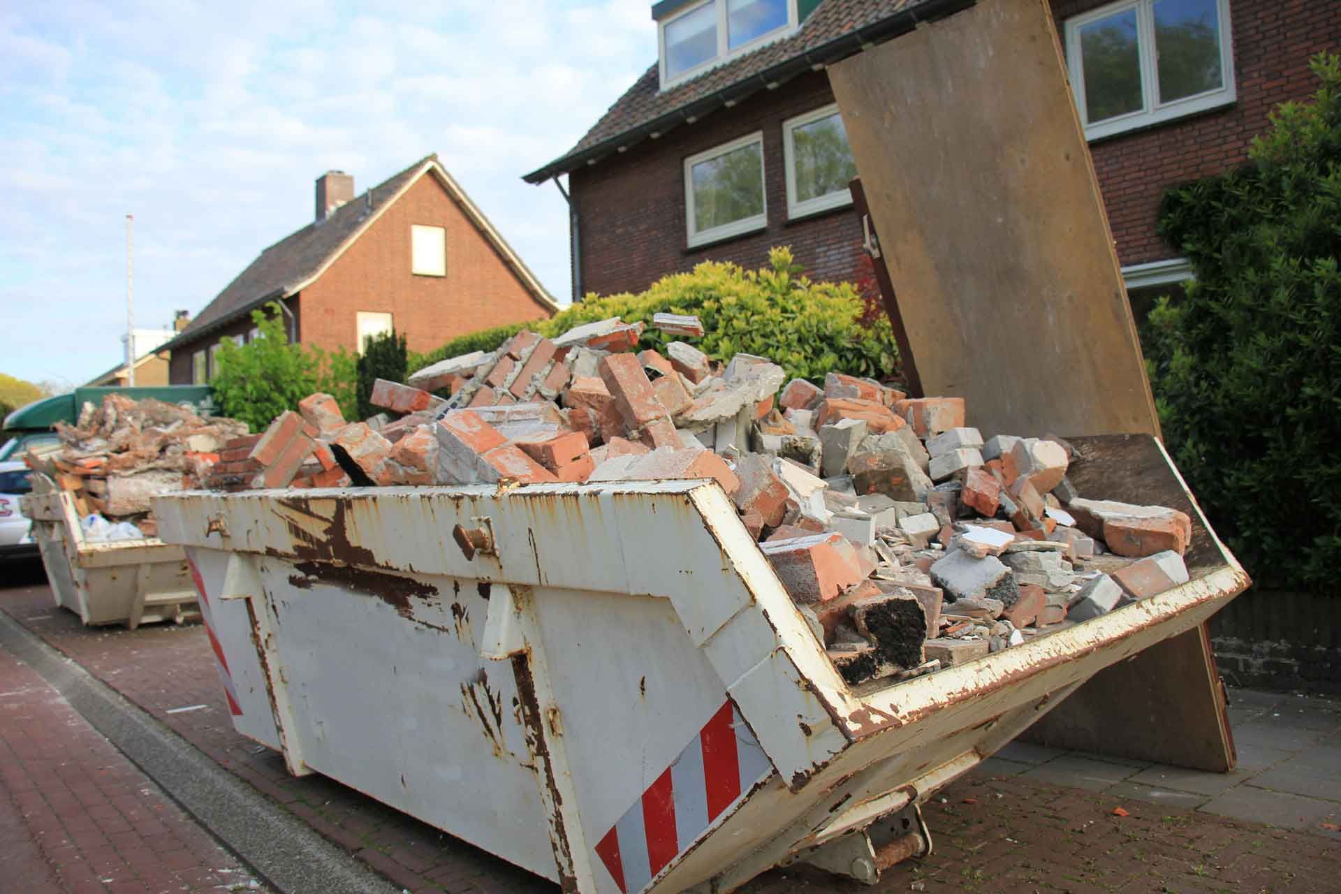 A metal dumpster overflowing with broken bricks and debris, likely from a construction site, in front of a residential building.