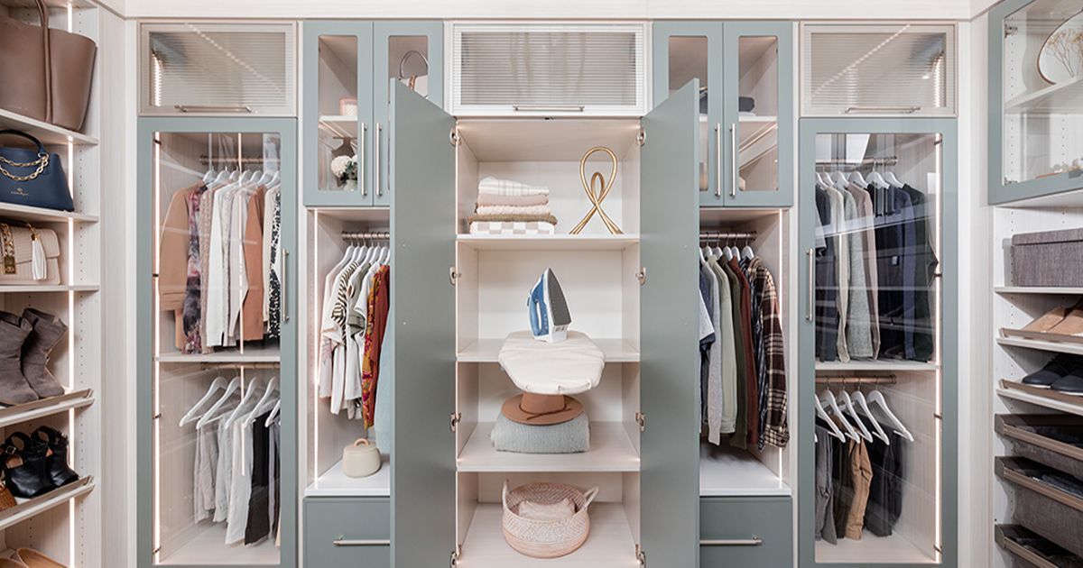 A modern, organized walk-in closet featuring blue-grey cabinetry, glass-front doors, hanging clothes, and an ironing board.
