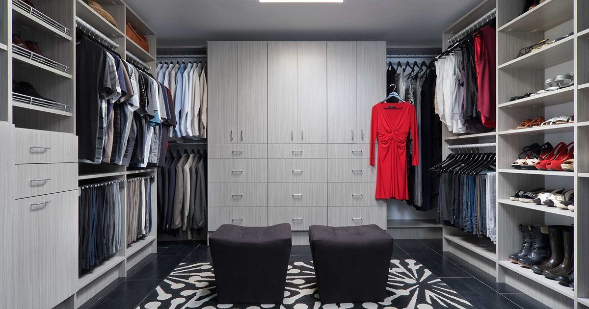 Walk-in closet with gray cabinets, hanging clothes, shelves of shoes, and two black ottomans on a patterned rug.