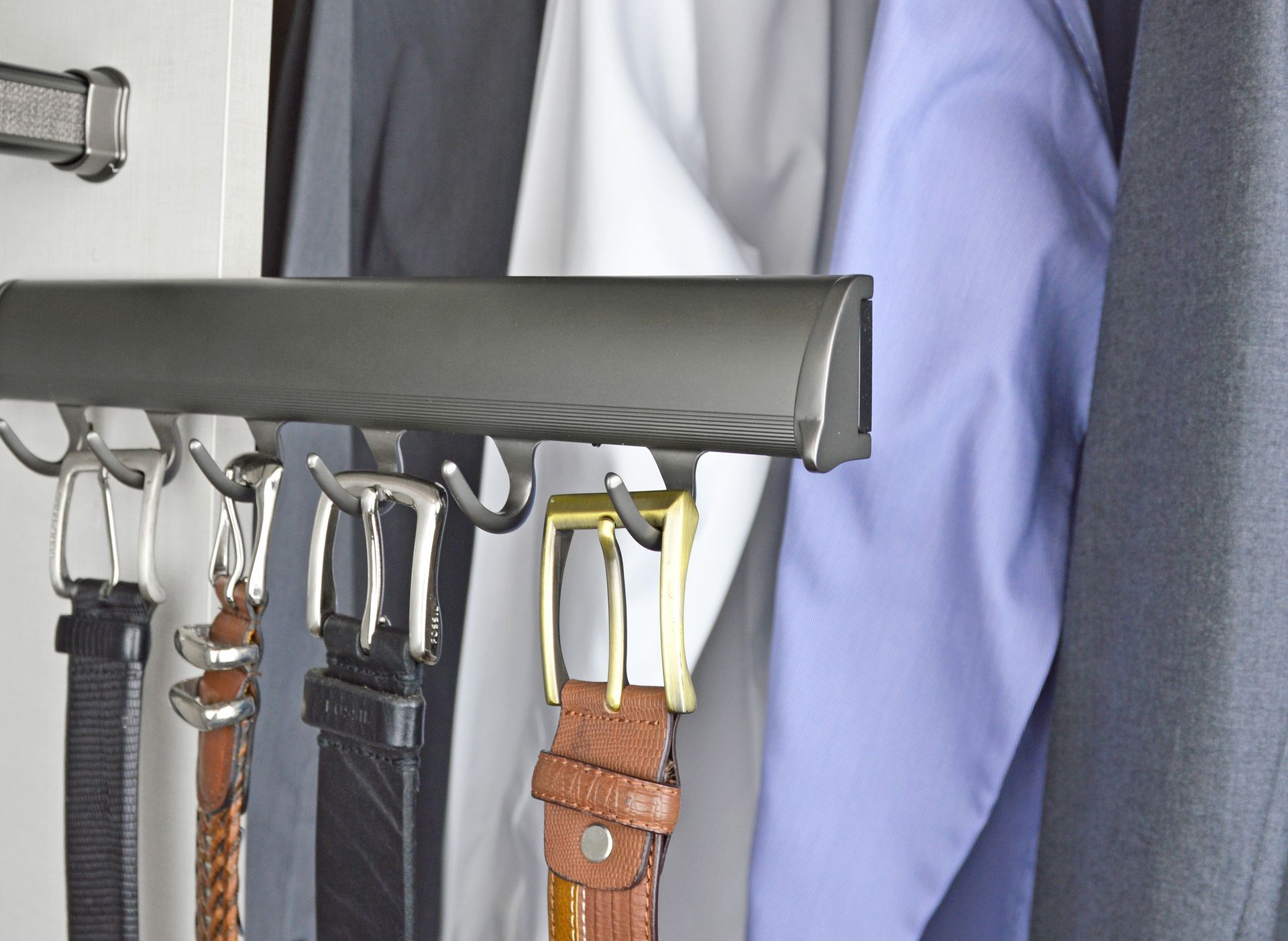 A dark gray sliding rack mounted in a closet, holding several belts with various metal buckles hanging from hooks.